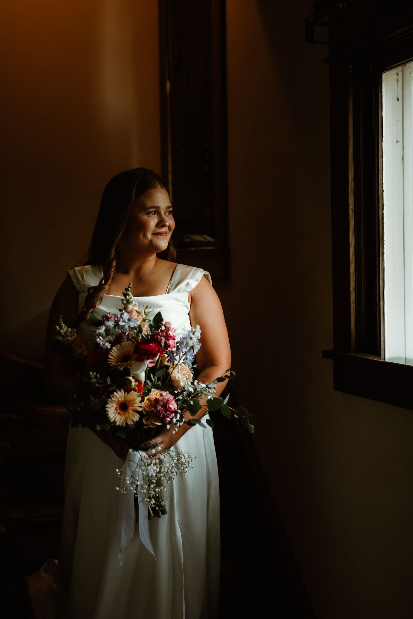 Bride in white gown holding colorful bouquet stands by window in soft natural light, smiling warmly.
