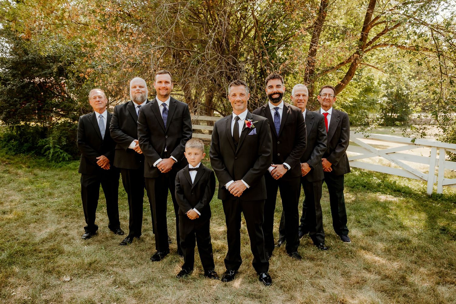 Groom stands with groomsmen and young ring bearer in black tuxedos outdoors among autumn trees for wedding party photo.