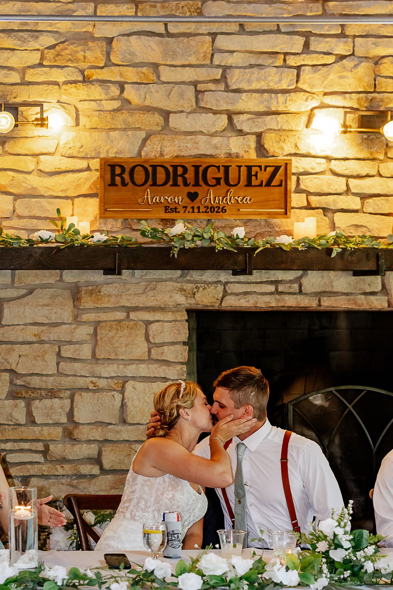 Newlyweds Aaron and Andrea Rodriguez share a kiss at their wedding reception in front of a stone fireplace, Est. 7.11.2026.
