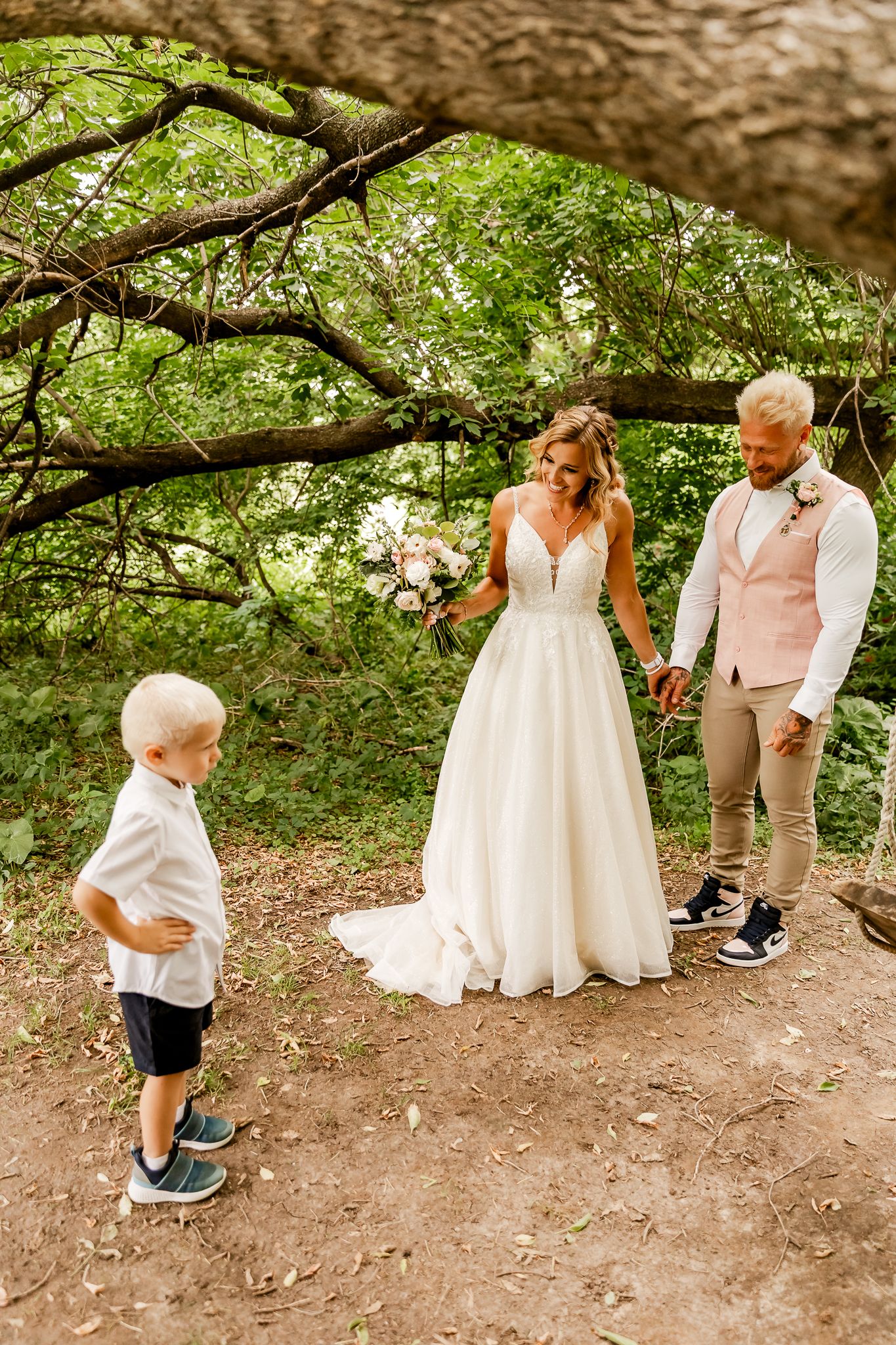 Bride and groom hold hands in a wooded setting while a young boy in wedding attire looks on.
