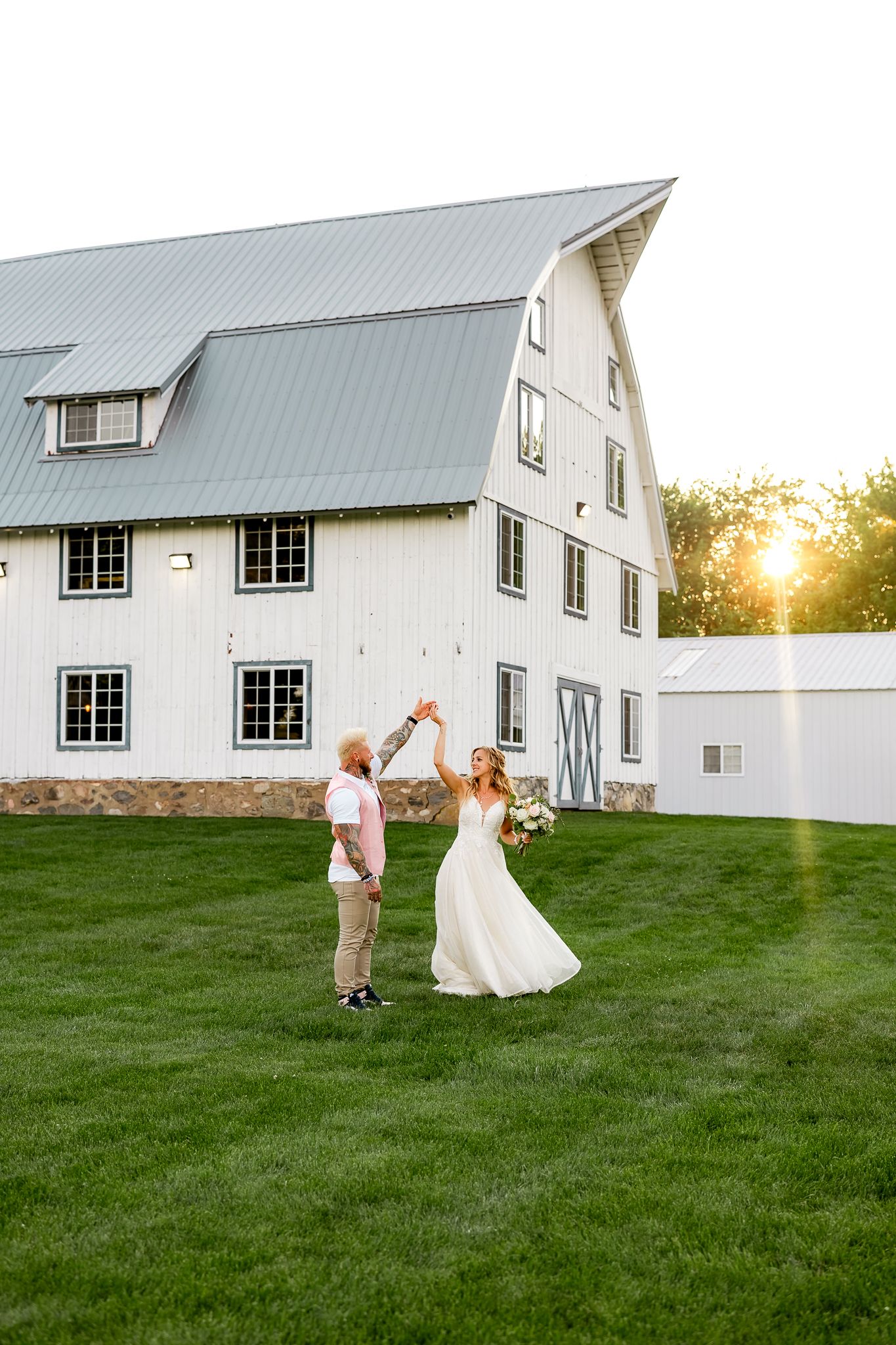 Bride and groom dancing on green lawn in front of a white barn venue at golden hour sunset.