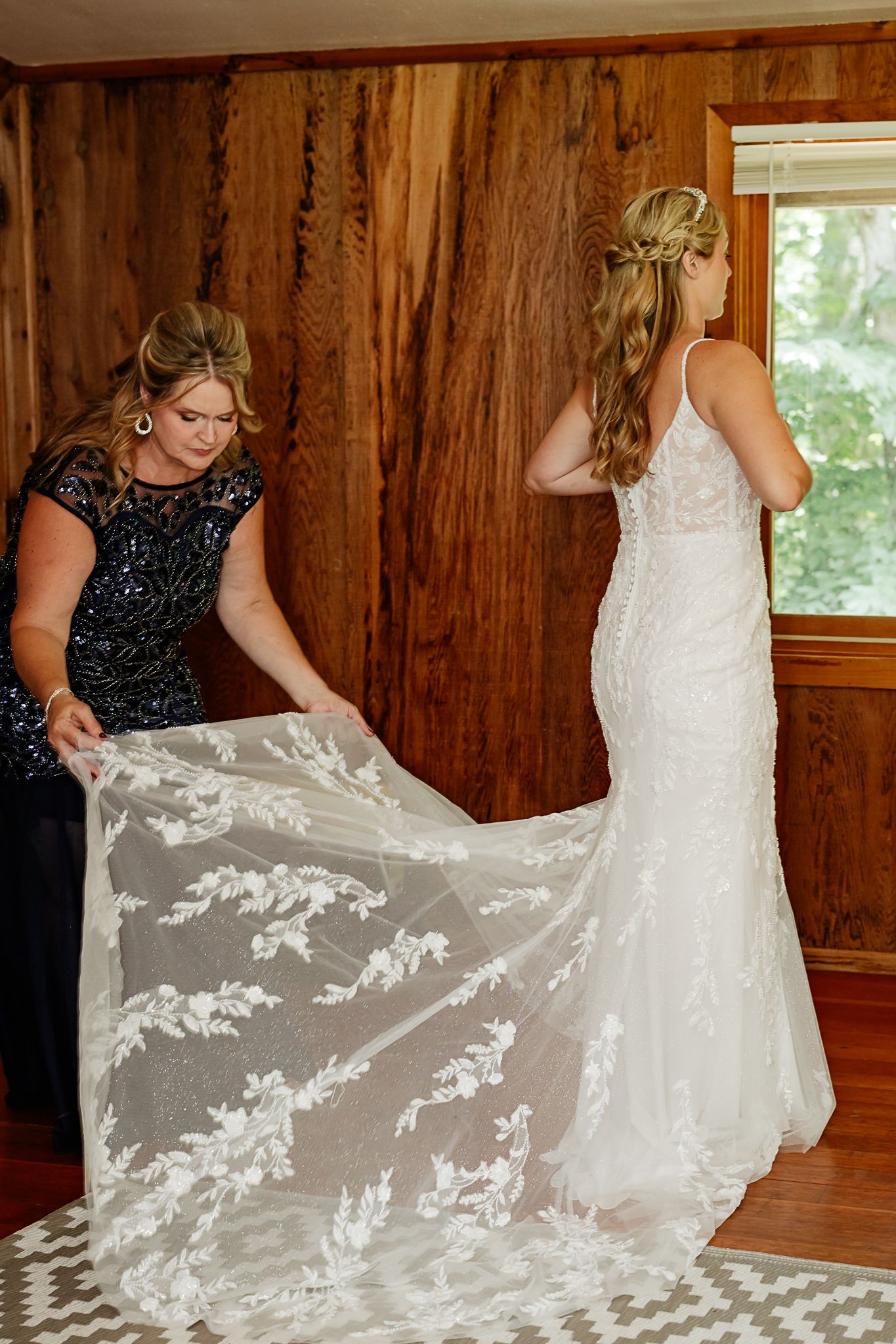 A bride in a lace wedding gown has her train adjusted by a woman in a dark beaded dress in a wood-paneled room.