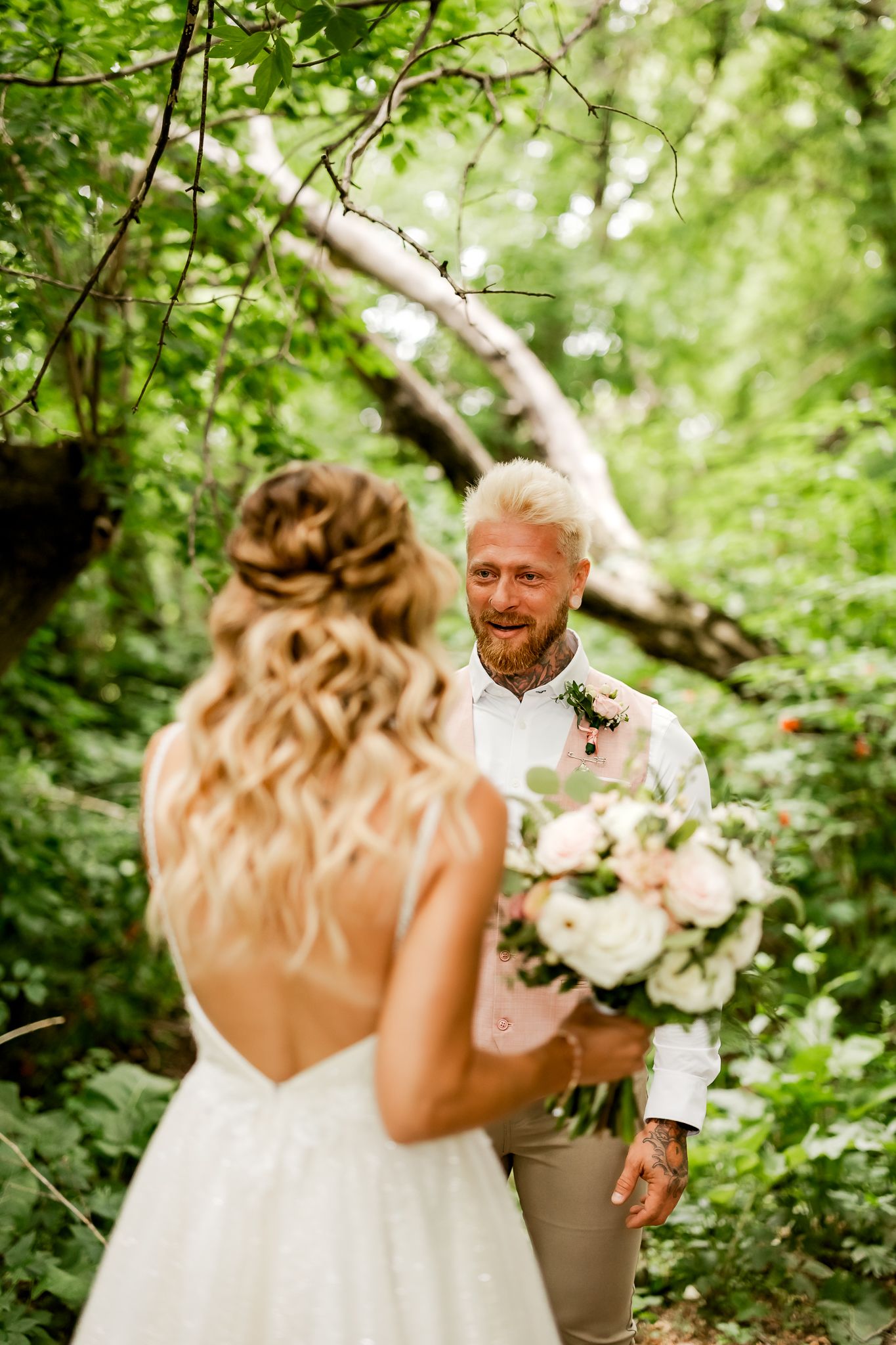 Smiling groom with beard and tattoos reacts emotionally seeing bride in white dress during outdoor forest wedding first look.