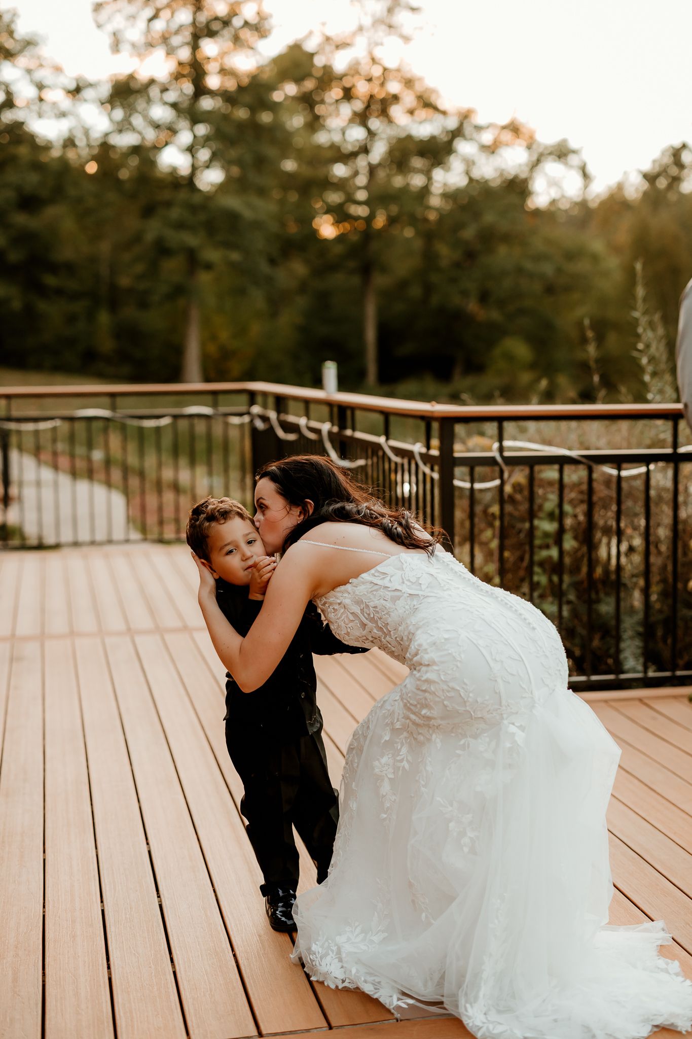 A bride in a lace wedding gown bends down to kiss a young boy in a black suit on a wooden deck at sunset.