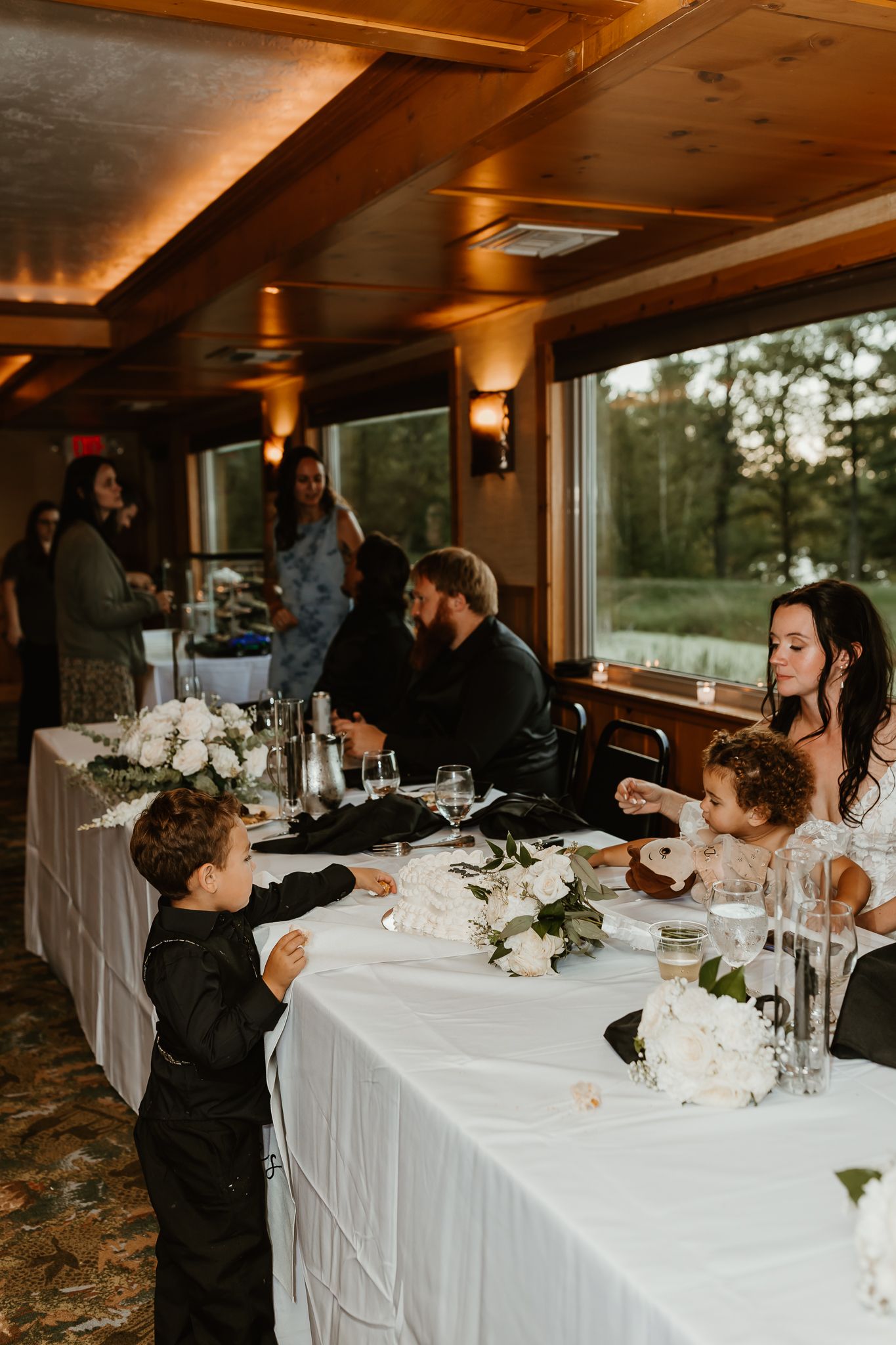 Wedding reception head table with white floral centerpieces, a boy in a black suit standing nearby, and guests seated by large windows.