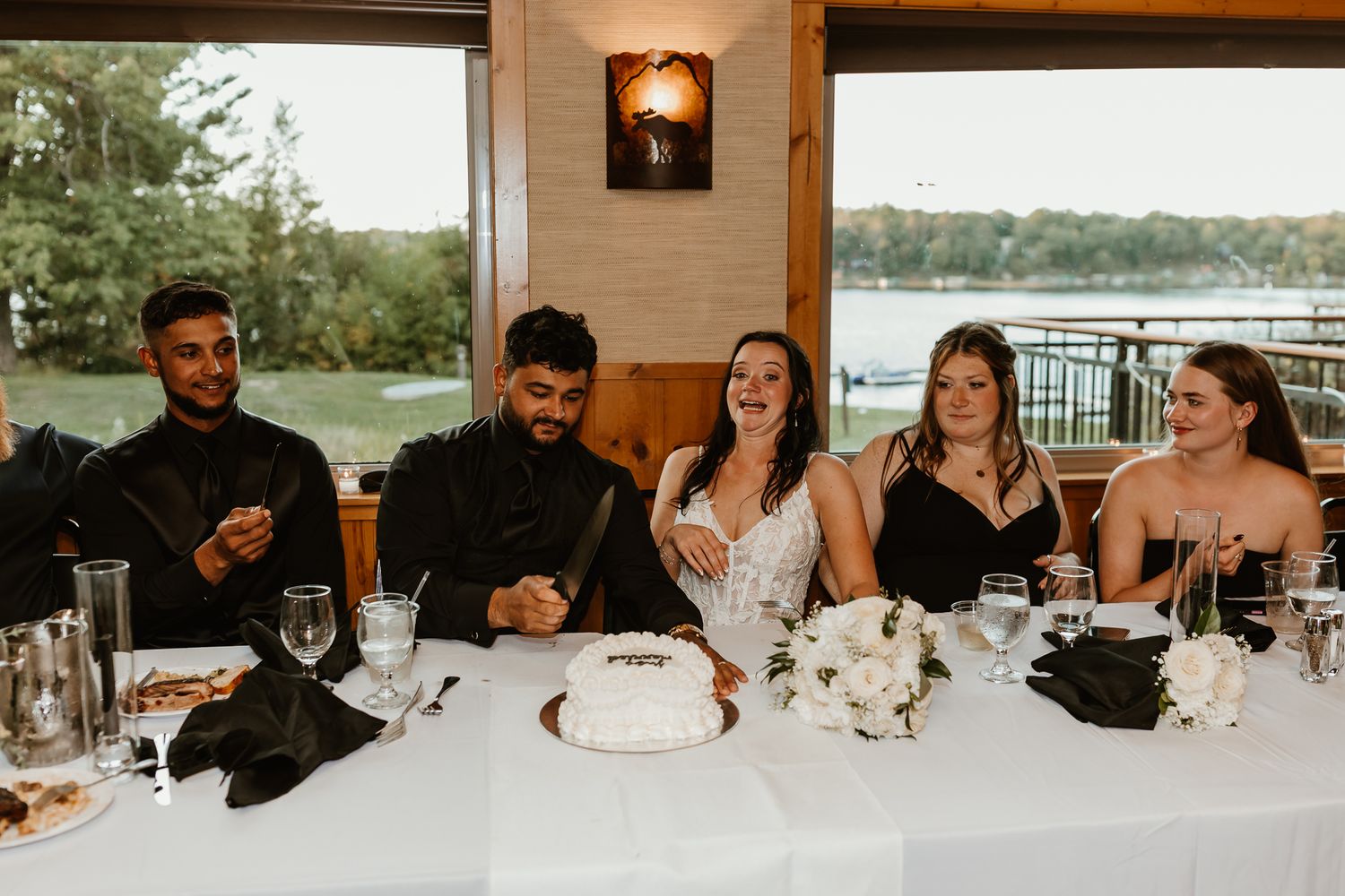 Wedding party seated at a decorated head table with a white cake and floral centerpiece, overlooking a scenic lake through large windows.