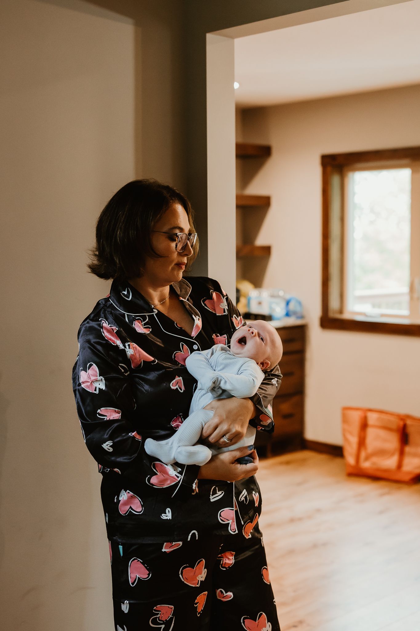 Woman in black heart-print satin pajamas gently holds a yawning newborn baby in a bright, warm room with wood floors and a window.