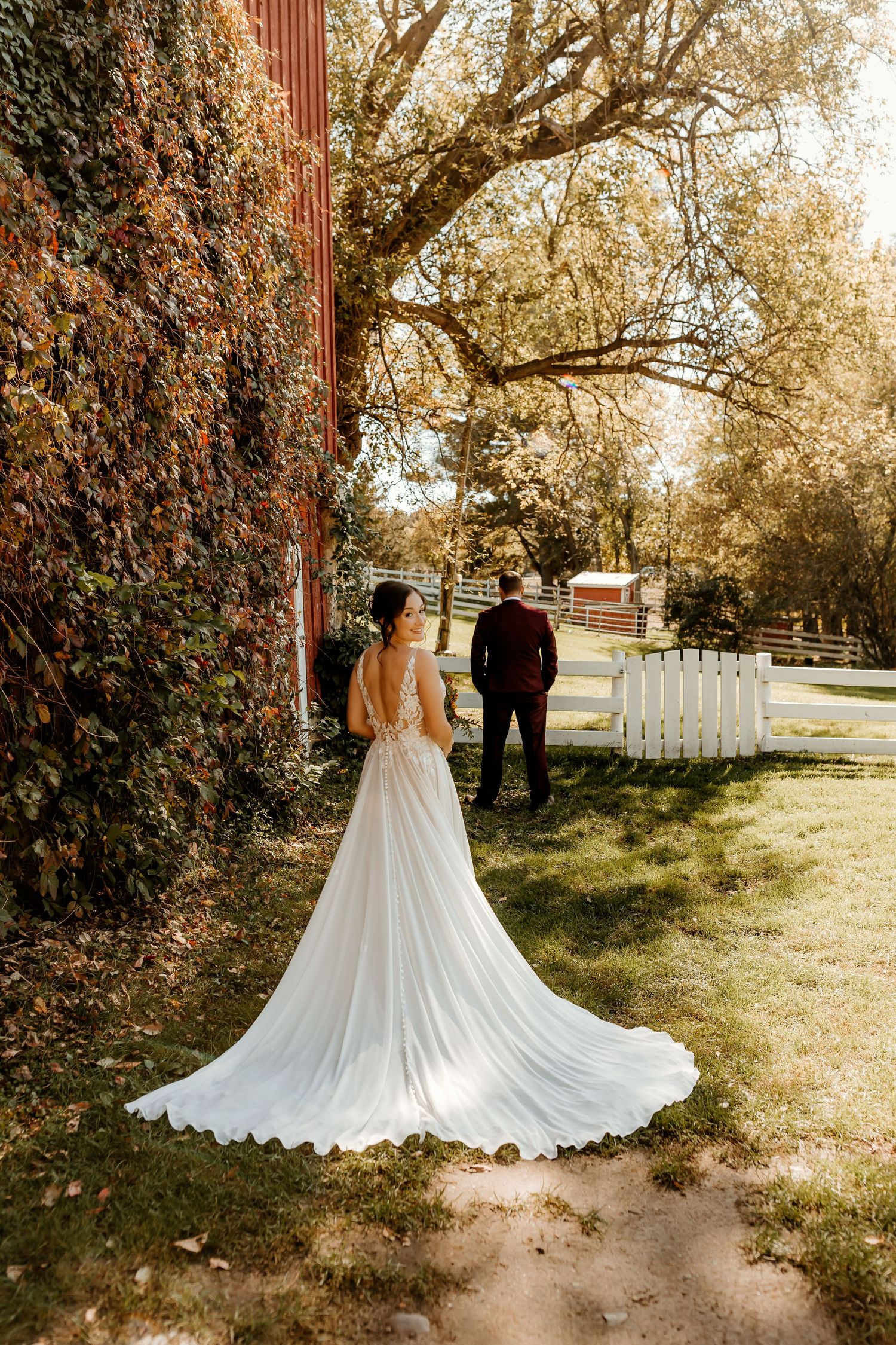 Bride in a flowing lace wedding gown with a long train stands on grass facing a groom in a burgundy suit near a red barn and white fence.
