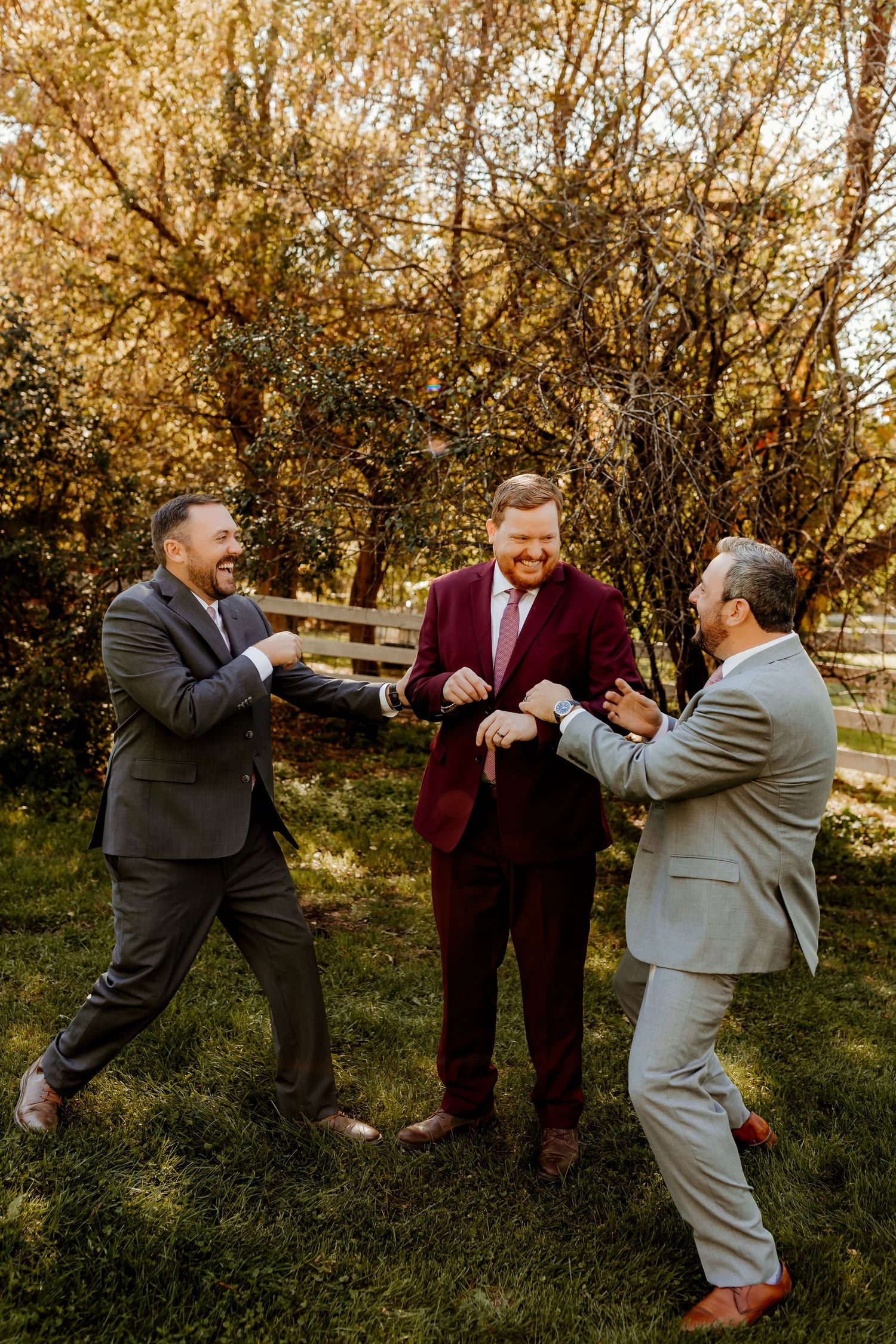 Three laughing men in gray and burgundy suits joke around on a sunny, tree-lined lawn in autumn.