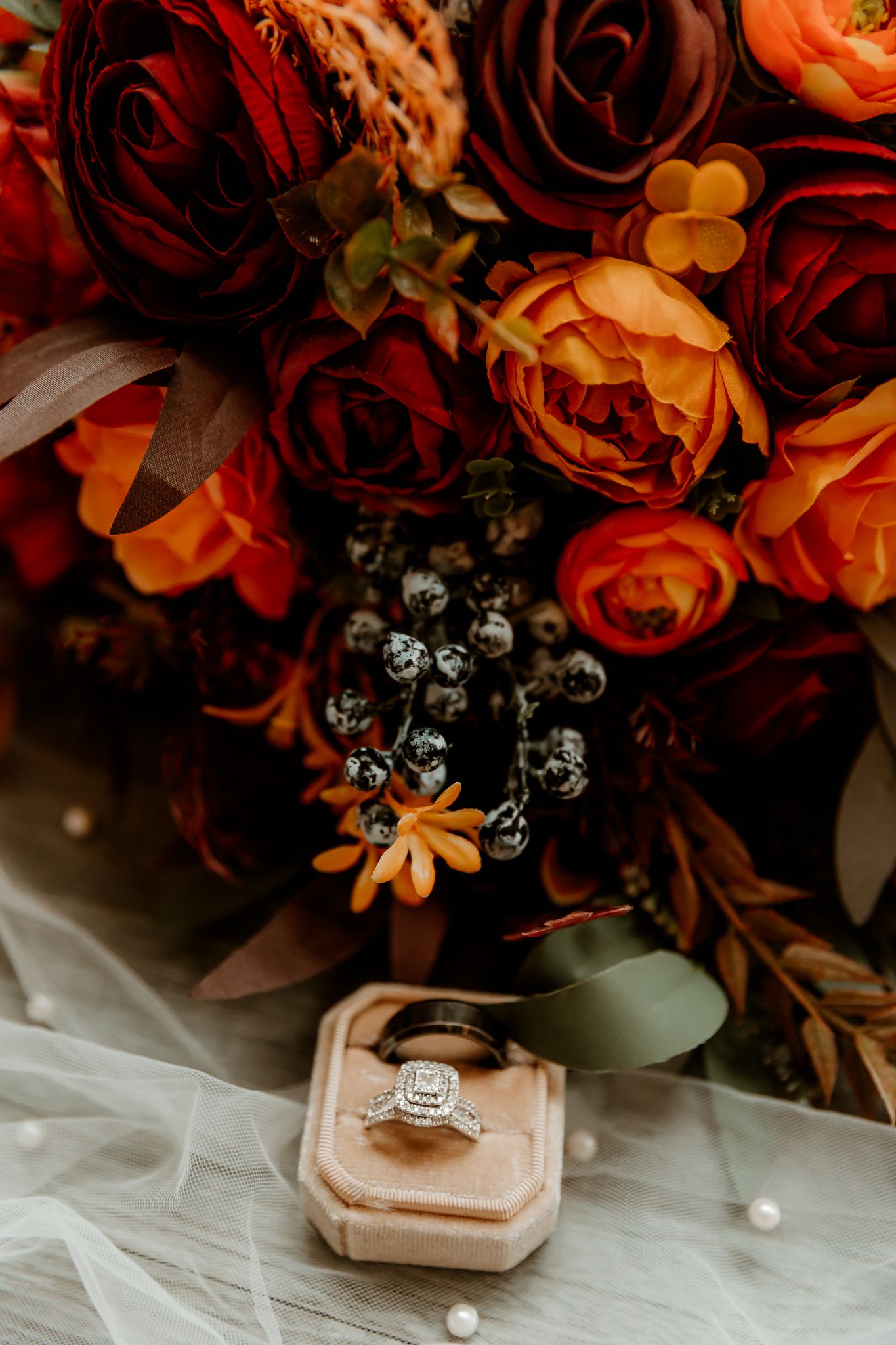 Burnt orange and burgundy wedding bouquet beside a diamond engagement ring and wedding band in a blush ring box on tulle.