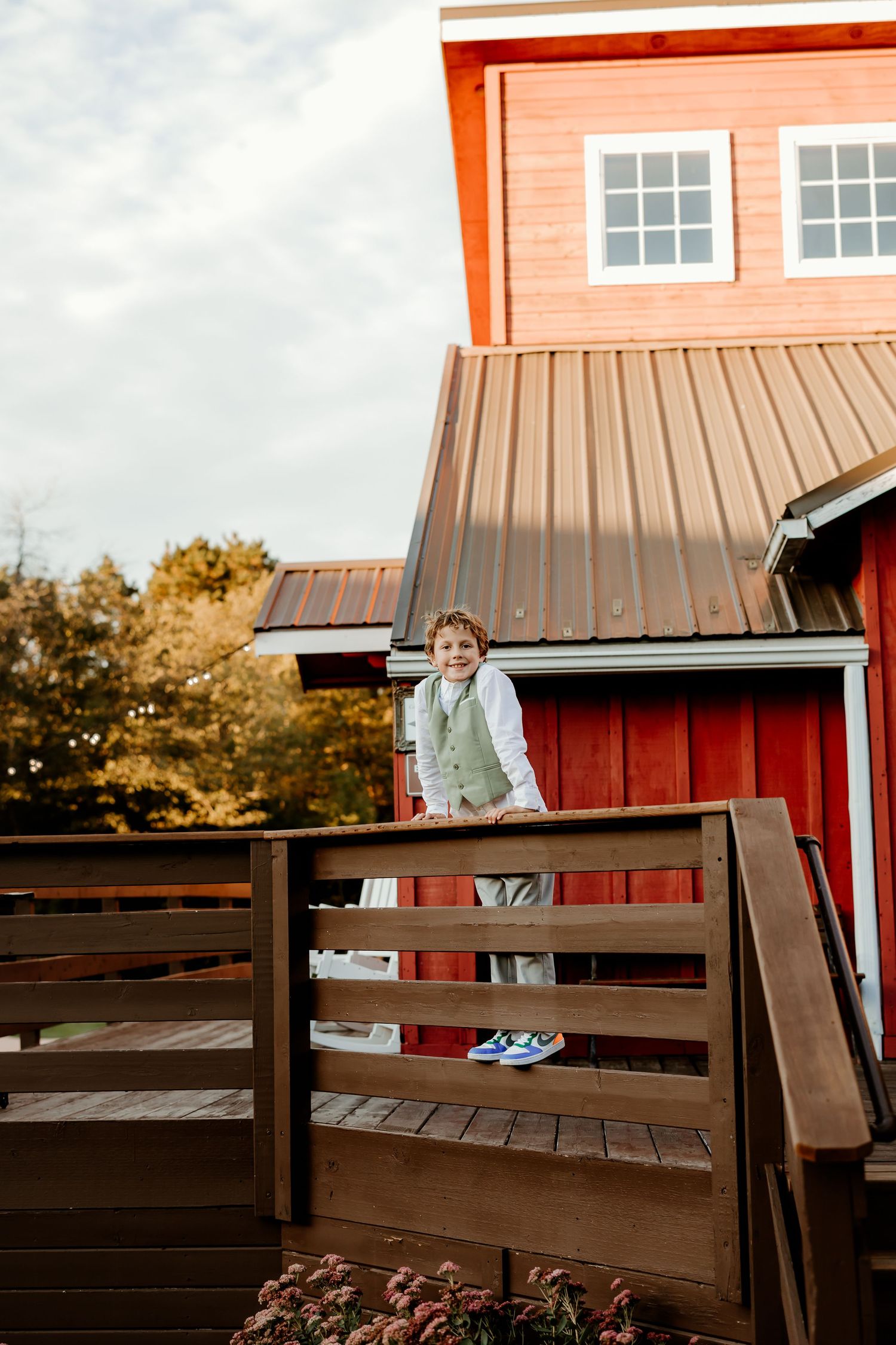 Smiling boy in a green vest and blue sneakers stands on a wooden deck railing in front of a red barn with a metal roof.