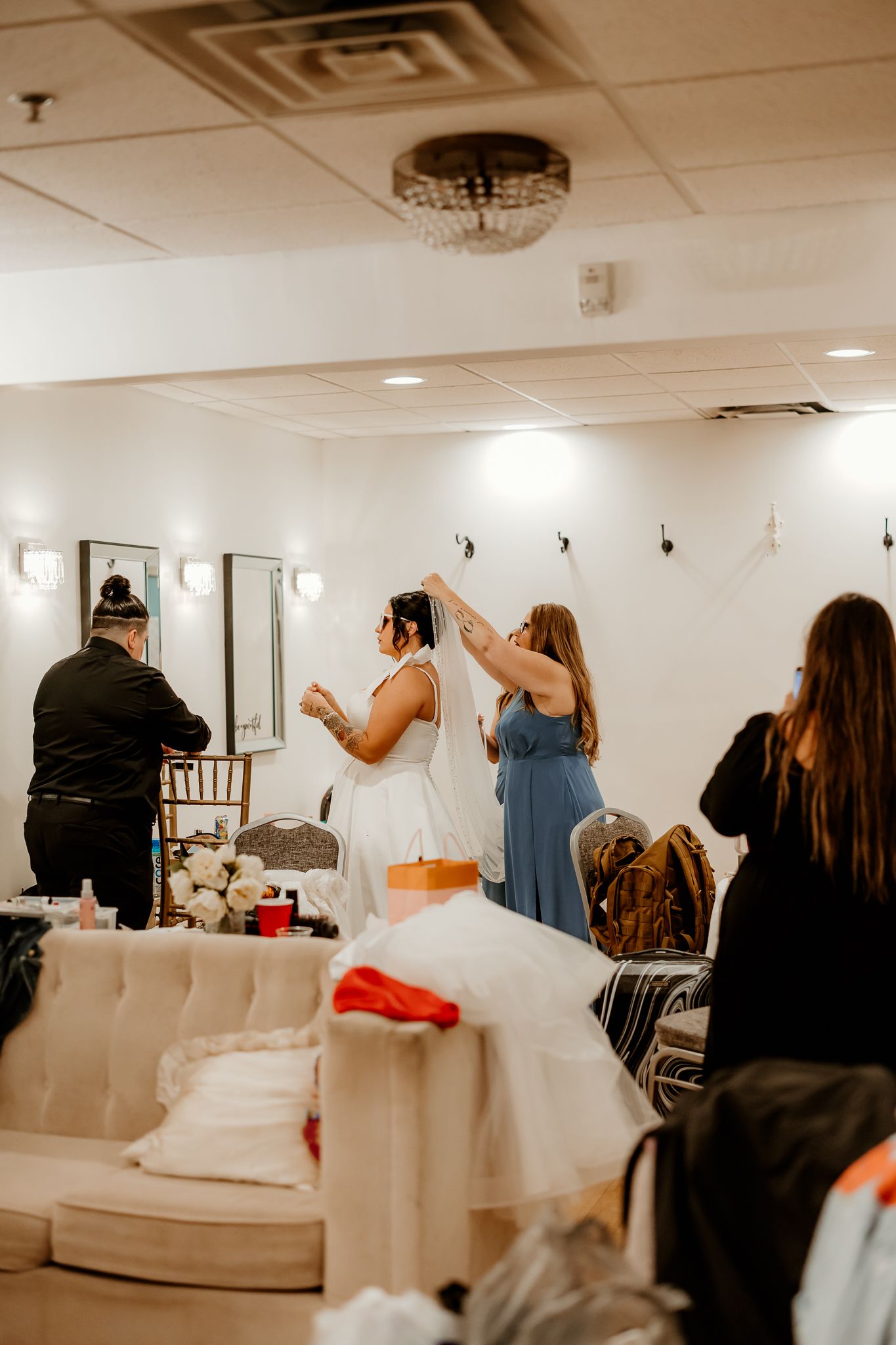 Bride in a white wedding dress has her veil adjusted by a bridesmaid in a blue dress in a bridal suite with a tufted sofa and vanity lights.