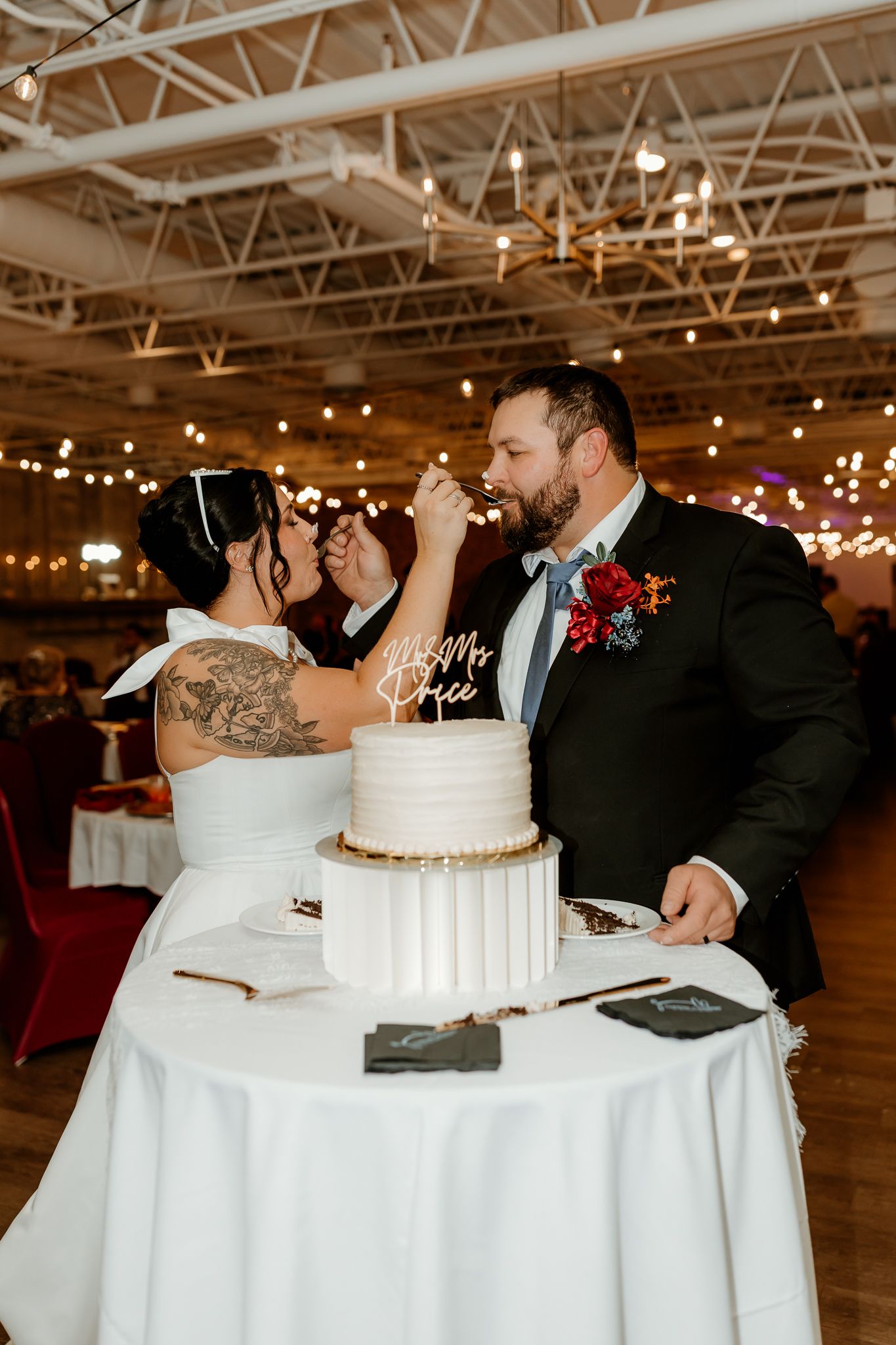 Bride and groom share a bite of their white tiered wedding cake at a reception with string lights and a red floral boutonniere.