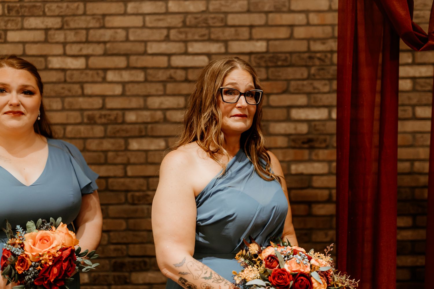 Two bridesmaids in dusty blue dresses hold orange and red floral bouquets while standing before a brick wall and burgundy curtain.