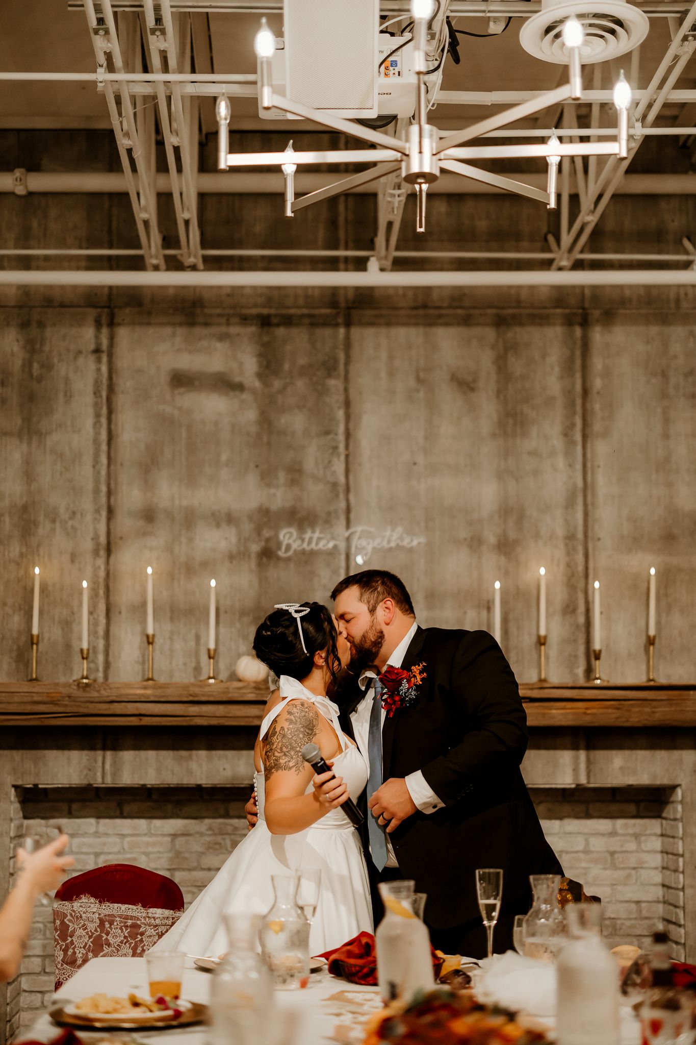Bride and groom share a kiss at their wedding reception in an industrial venue with candles, a modern chandelier, and a decorated table.