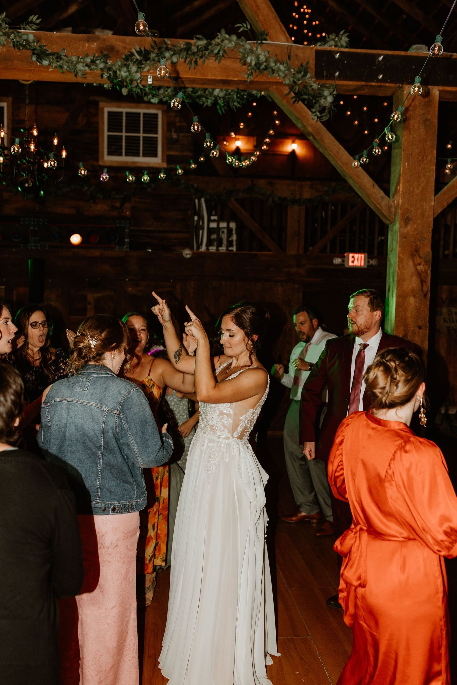 Bride in a flowing white lace gown dances with guests at a rustic barn wedding reception under string lights and greenery.