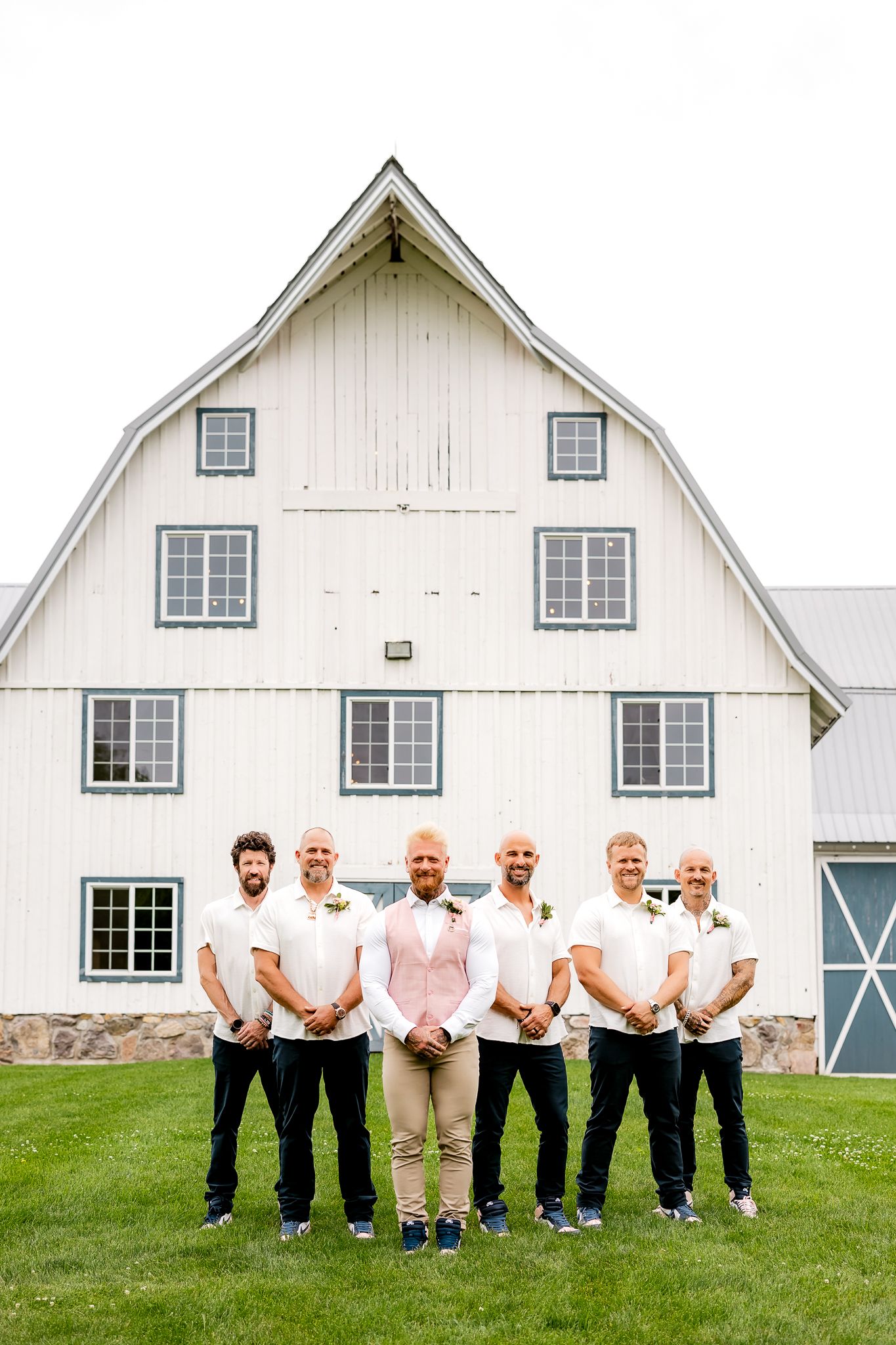 Groom in pink vest with groomsmen in white polos posing in front of a white barn at a rustic wedding.