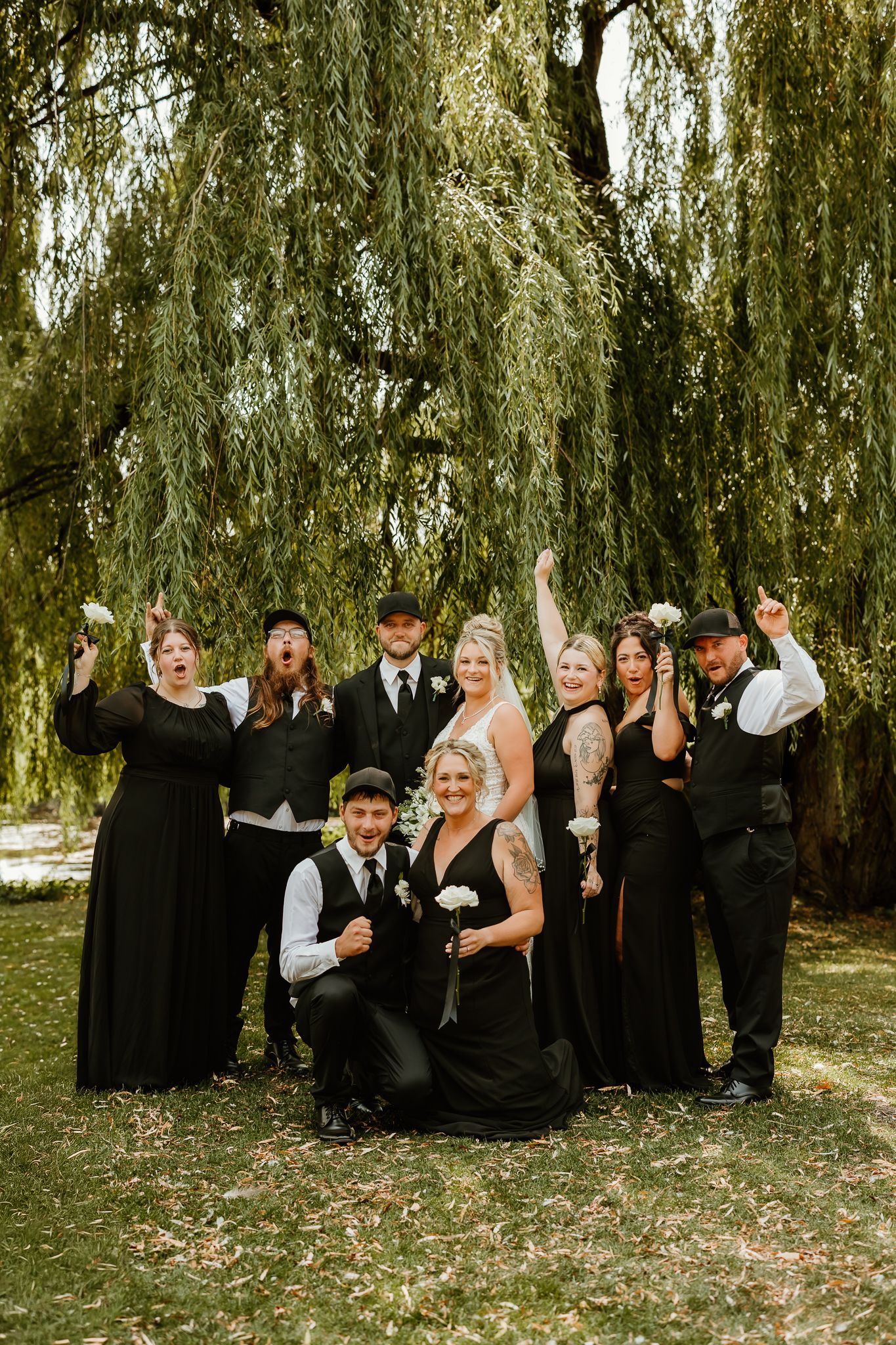 Wedding party in black attire celebrates joyfully under a weeping willow tree outdoors.