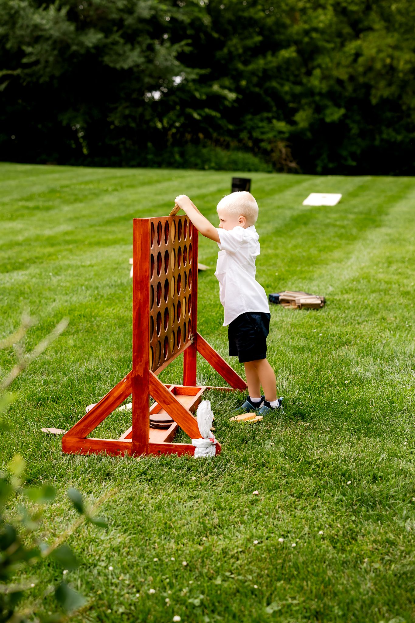 Young blonde boy playing giant Connect Four game on lush green lawn outdoors.
