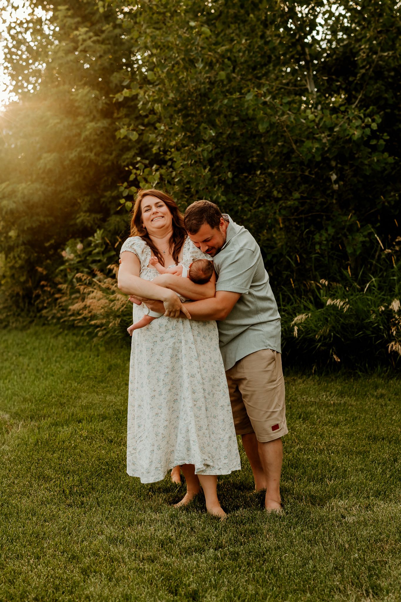 A joyful couple stands on green grass at golden hour, smiling as they hold their newborn baby together outdoors.