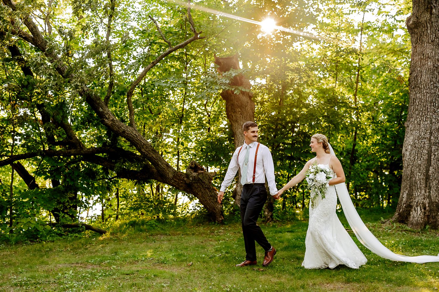 Bride and groom walk hand-in-hand through a sunlit green forest on their wedding day.