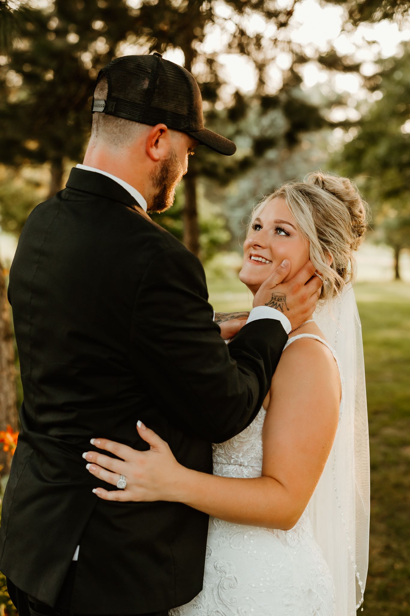A bride in a white lace gown smiles joyfully as her groom in a black suit and cap tenderly holds her face outdoors.
