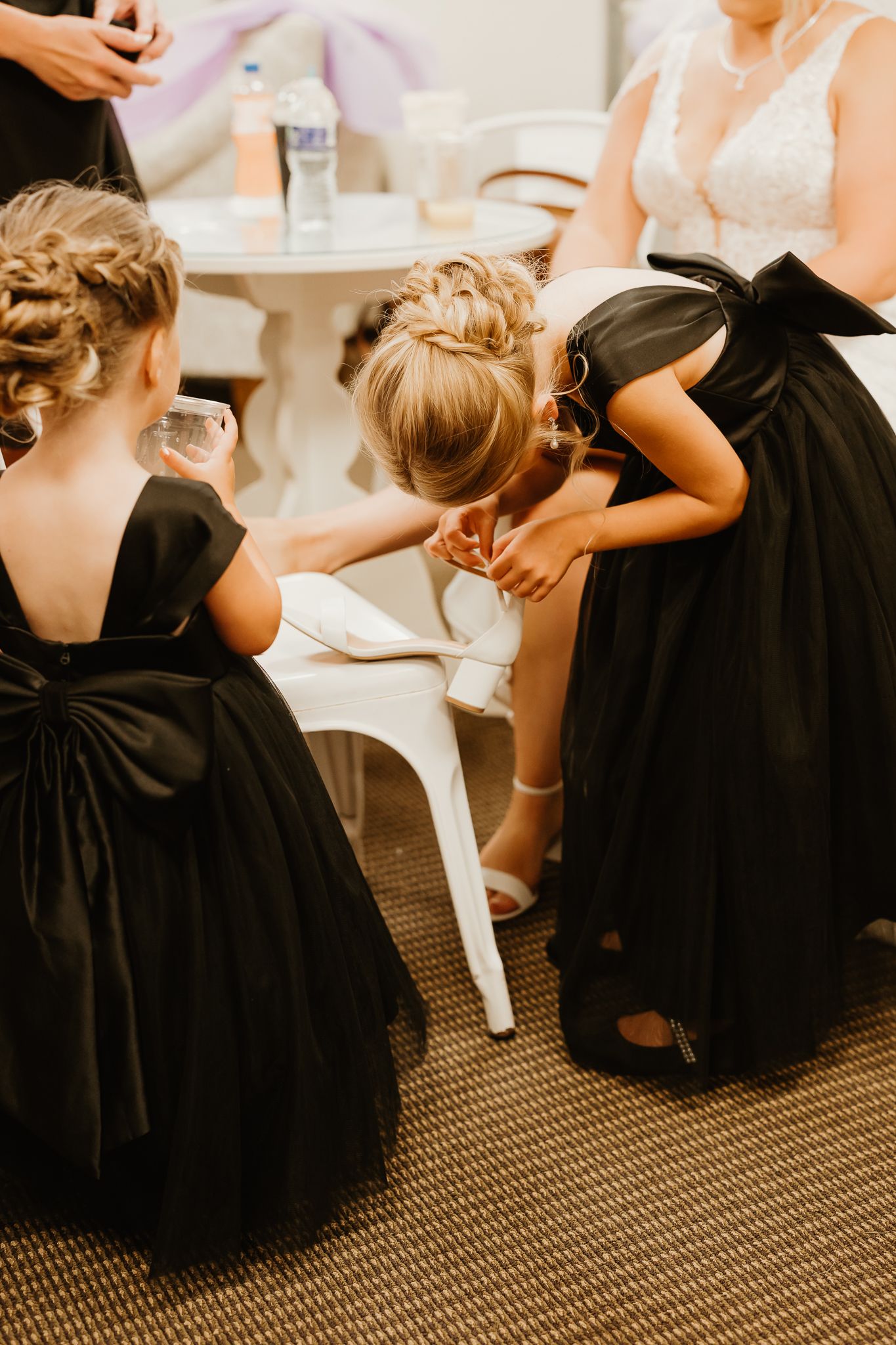 Two flower girls in black dresses with bows help bride with her shoes at wedding reception.