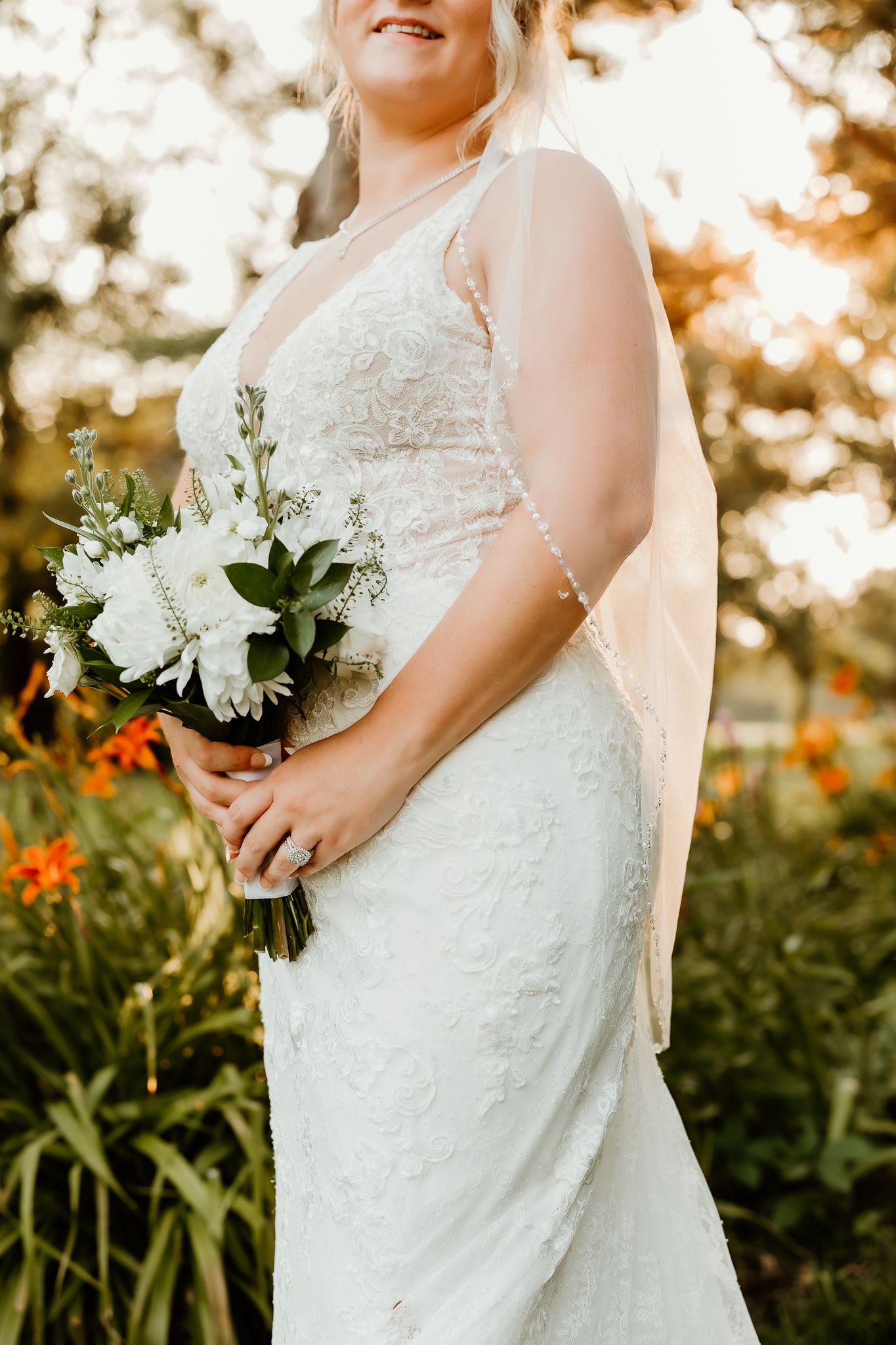 Bride in lace wedding gown holding white floral bouquet outdoors with golden hour sunlight and greenery background.
