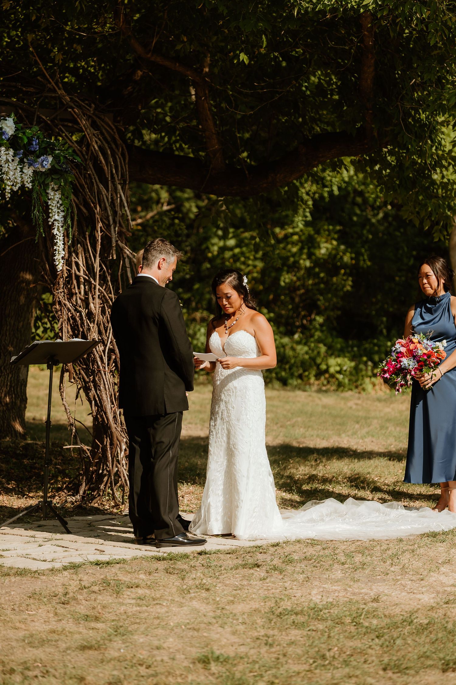 Bride reads vows to groom outdoors under a tree, with bridesmaid holding colorful bouquet during wedding ceremony.