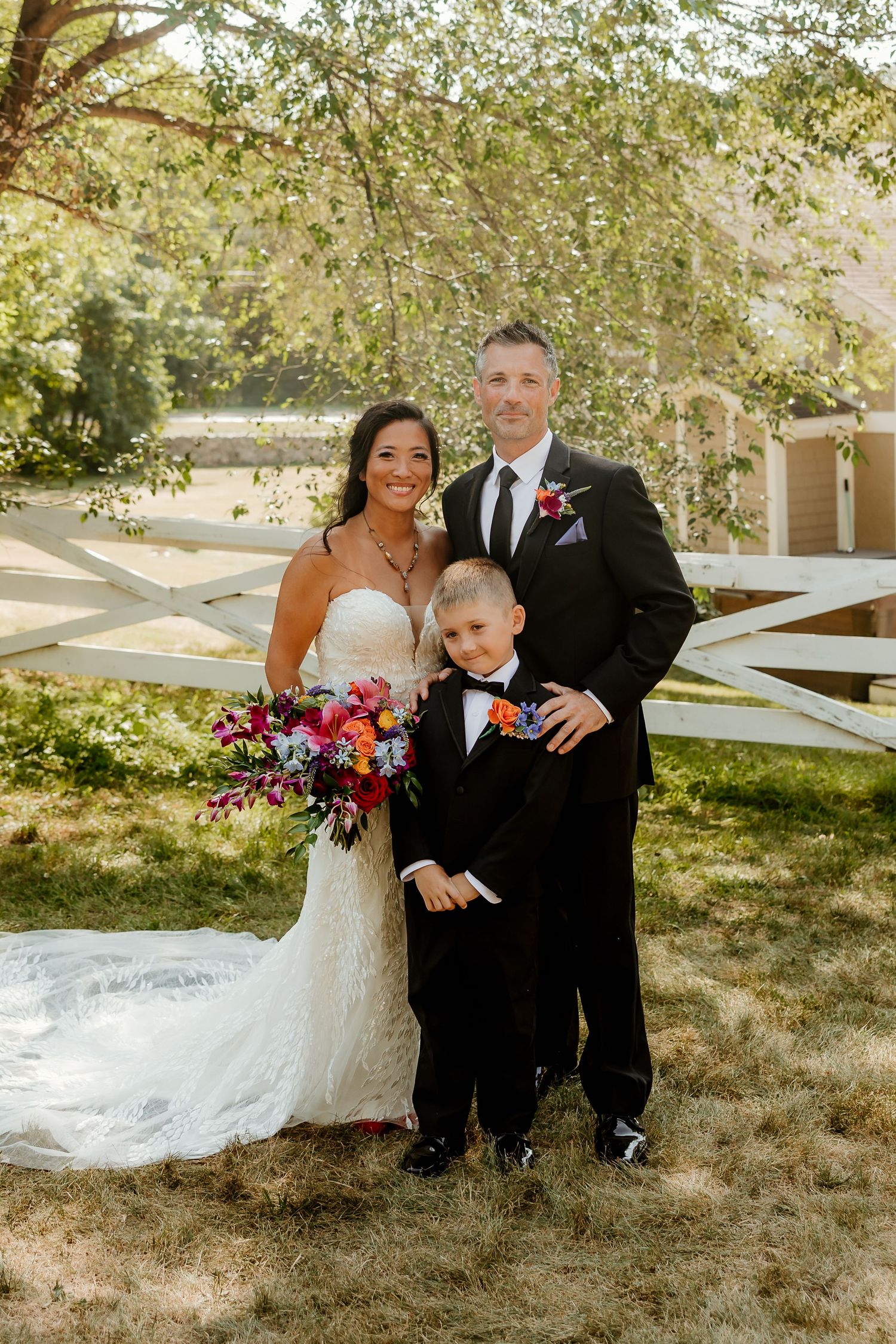 Bride and groom in formal wedding attire pose outdoors with young boy in tuxedo near white fence.