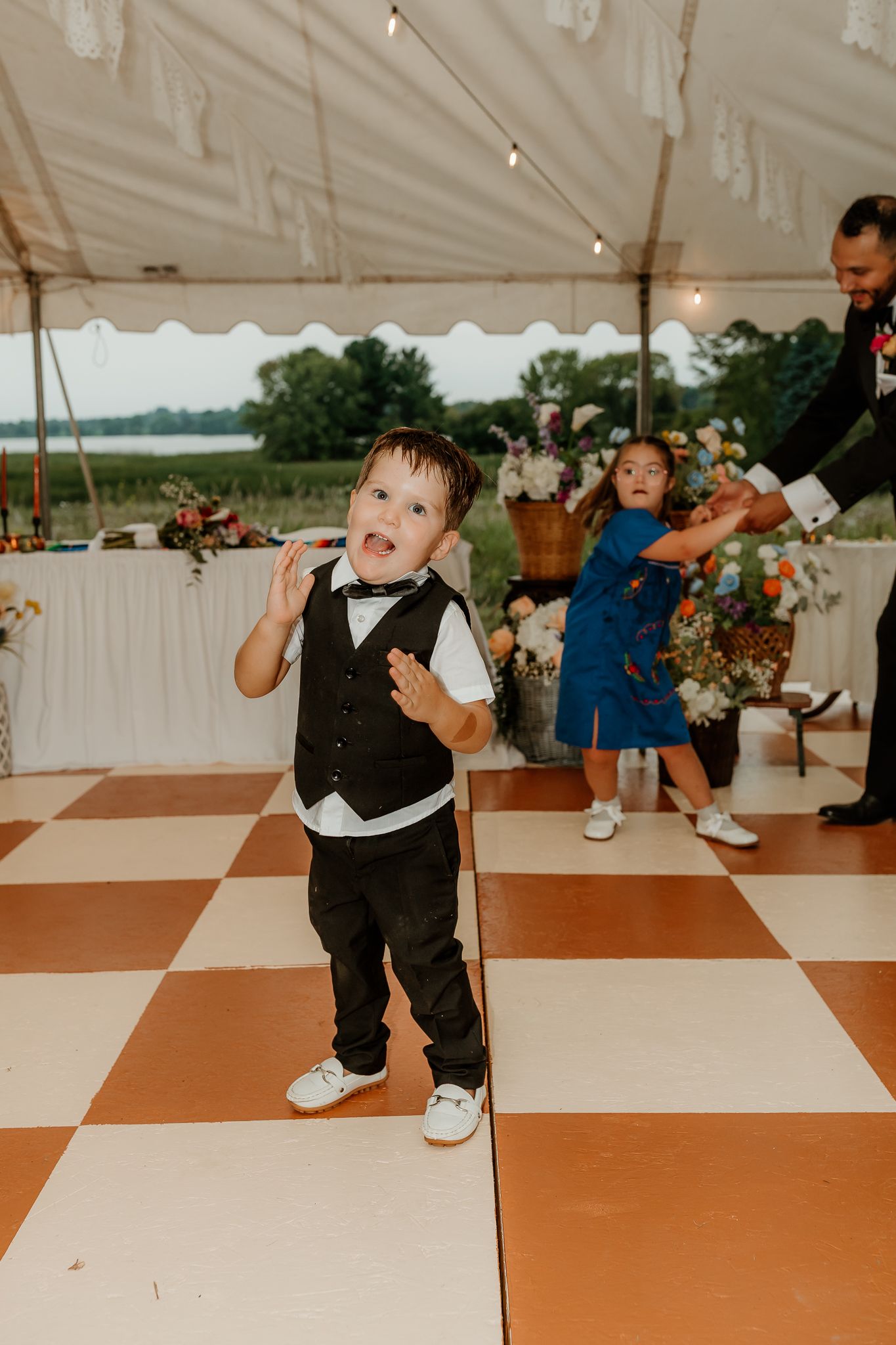 Young boy in black vest and bow tie dancing at a wedding reception on a checkered dance floor under a tent.