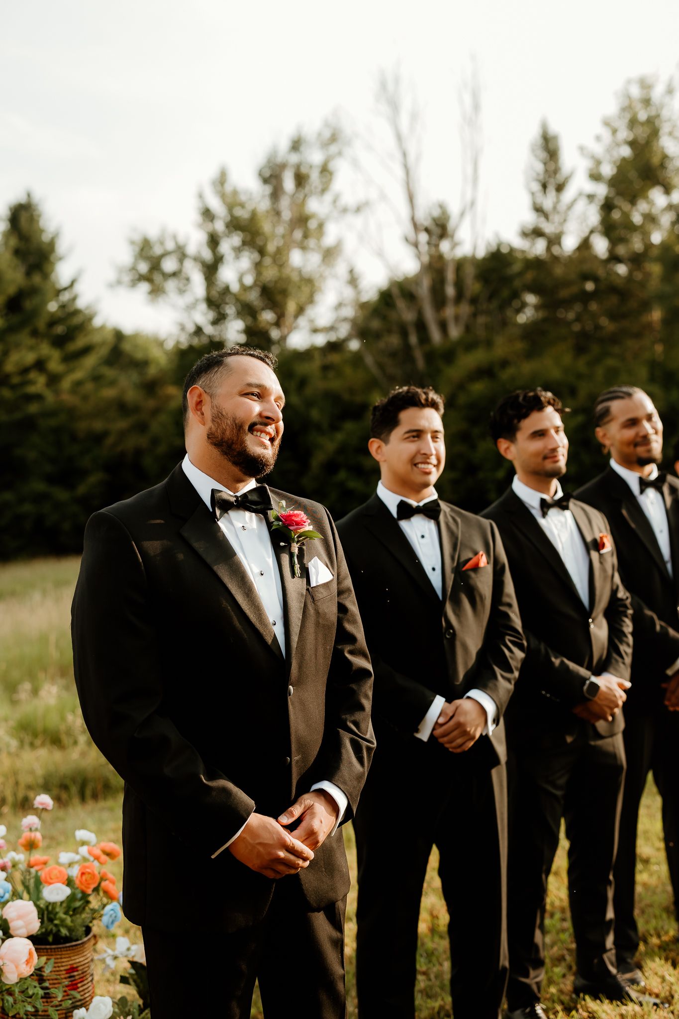 Groom and groomsmen in black tuxedos with boutonnieres stand outdoors at a wedding ceremony.