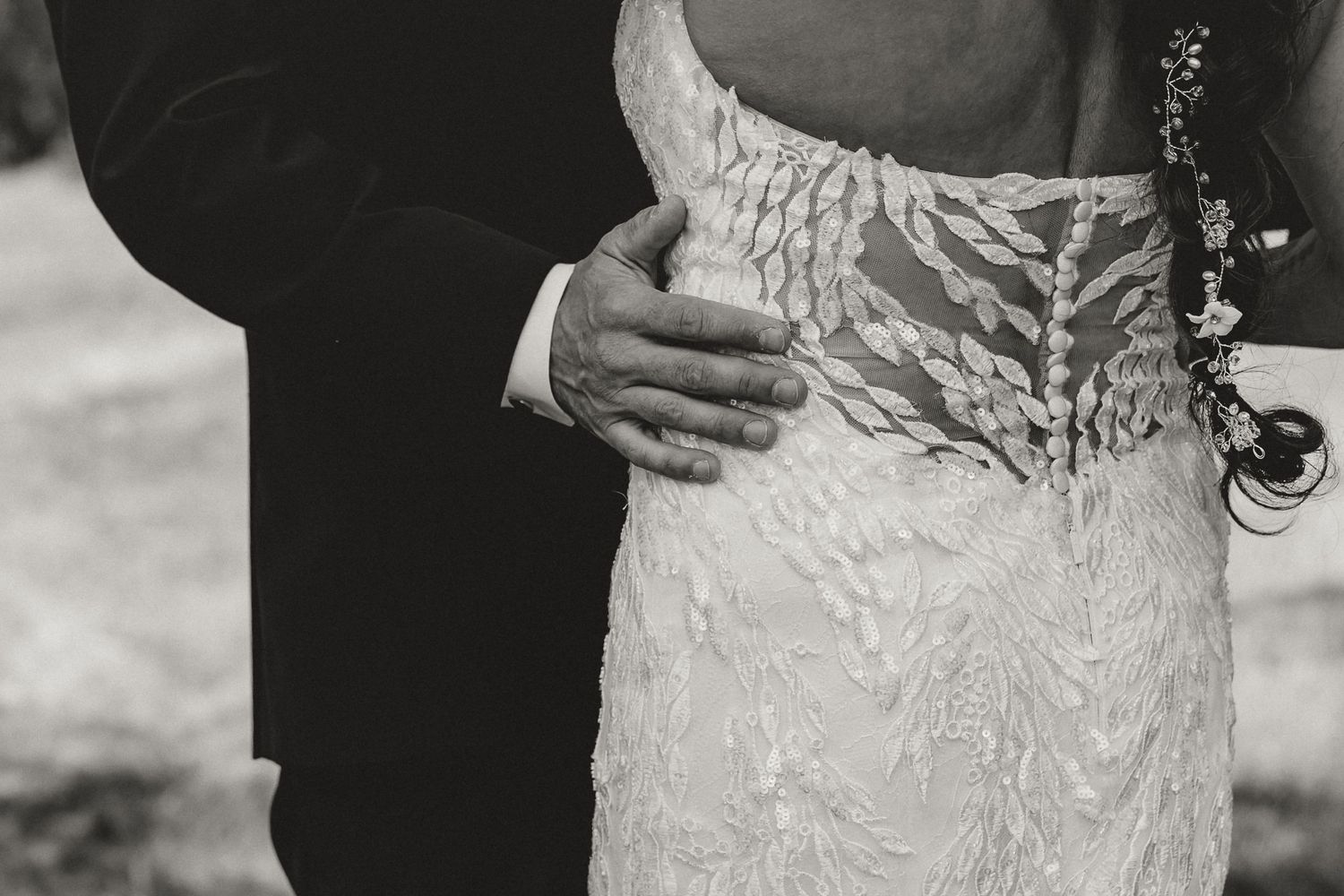 Black and white wedding photo of groom's hand resting on bride's lace dress waist.