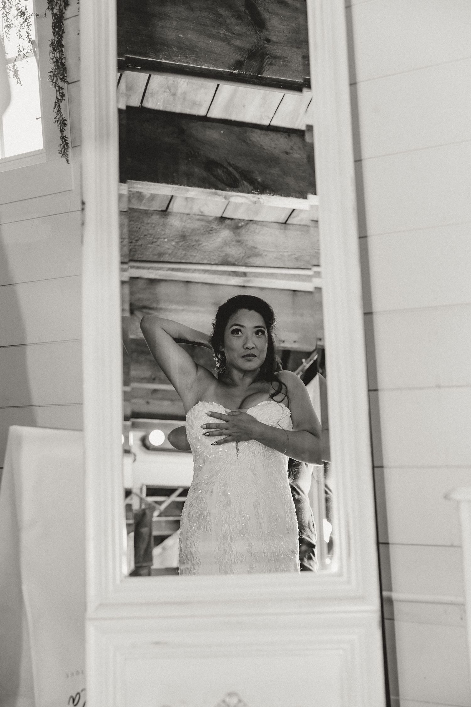Black and white photo of a bride in a lace gown adjusting her hair, reflected in a vintage mirror.