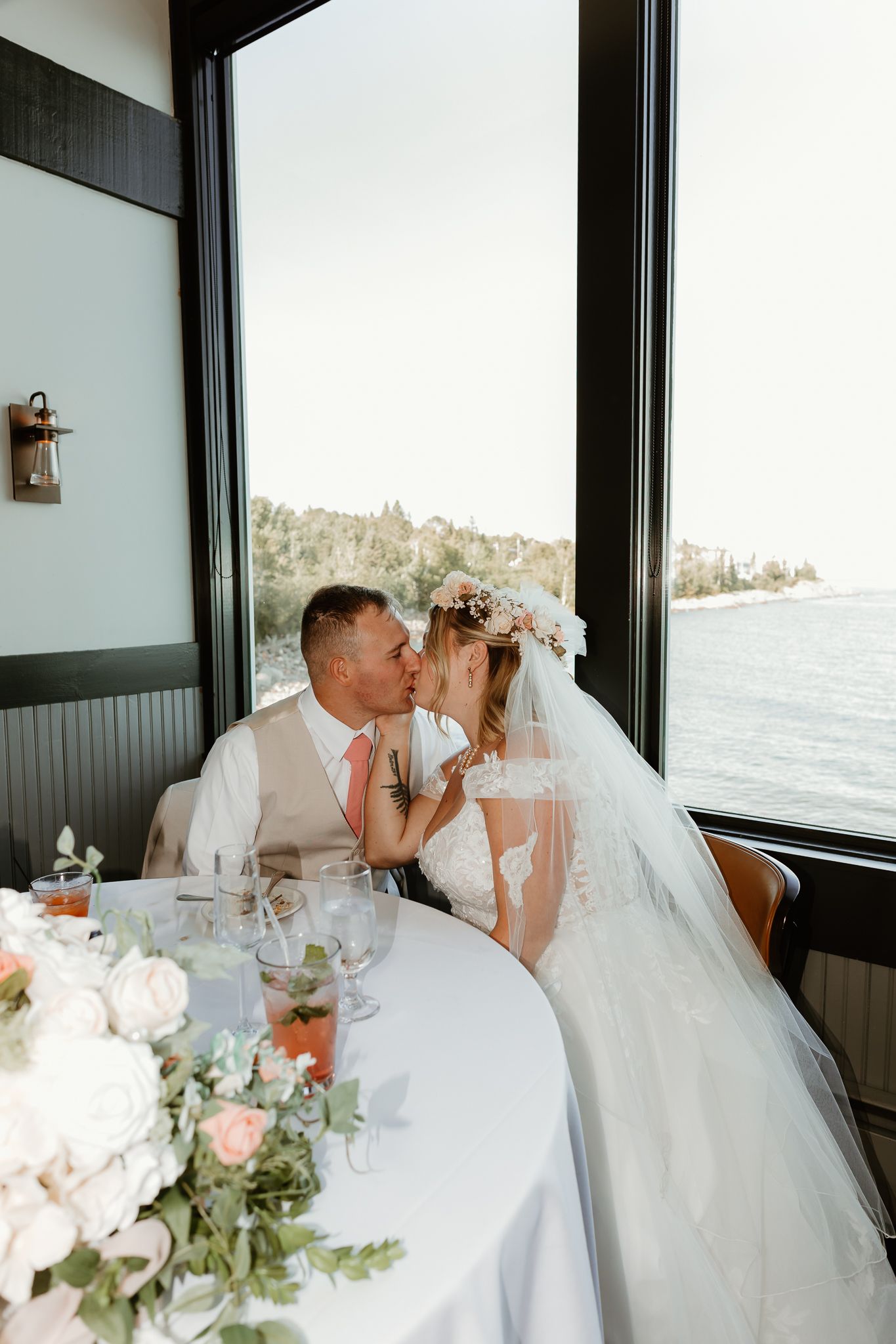 Bride and groom share a kiss at their waterfront wedding reception table adorned with white and pink floral arrangements.