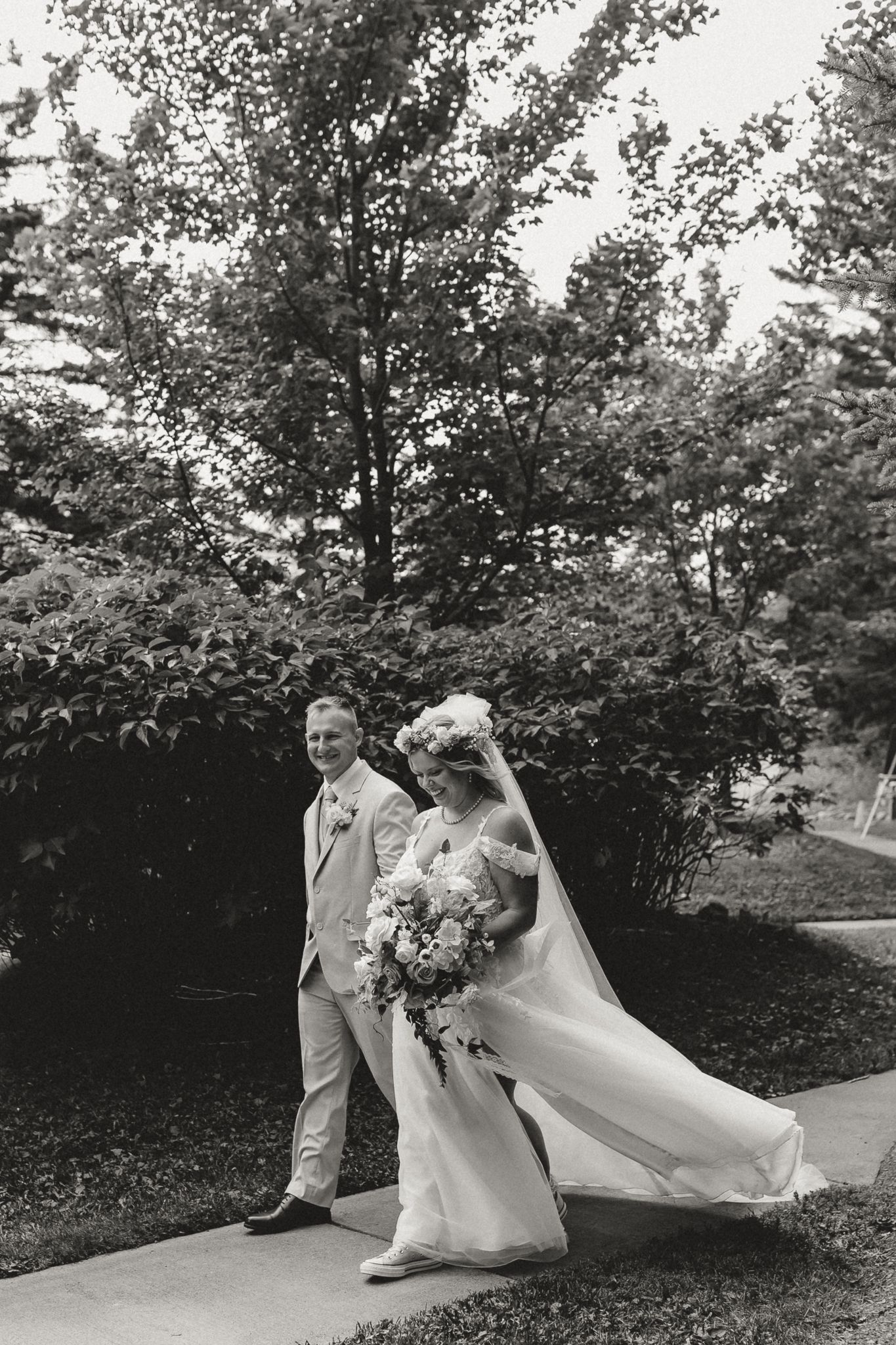 Black and white photo of a bride and groom walking outdoors, bride holding a large bouquet, surrounded by lush trees.