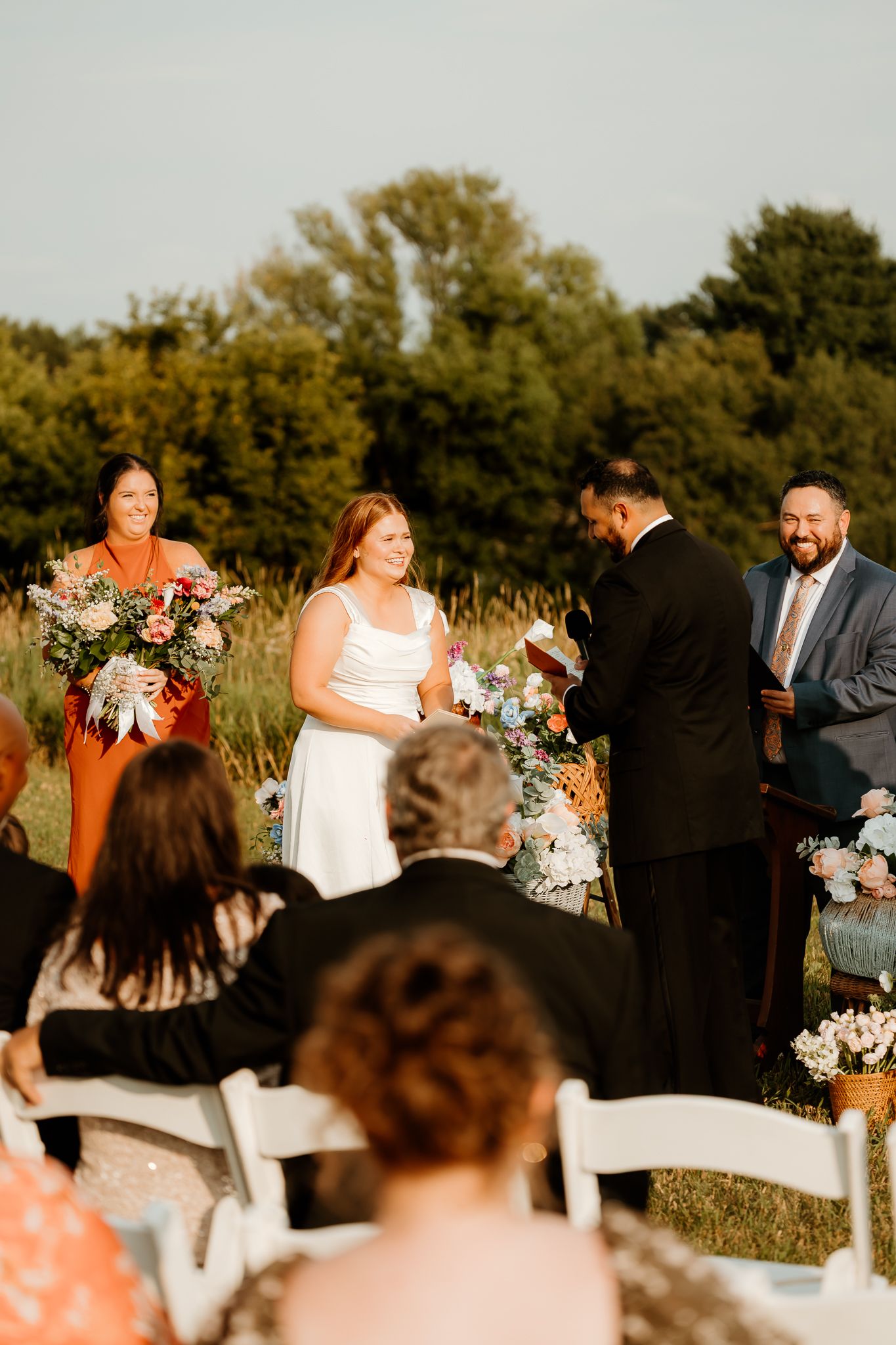 Outdoor wedding ceremony with bride in white dress, bridesmaid in orange, and officiant reading vows before seated guests.