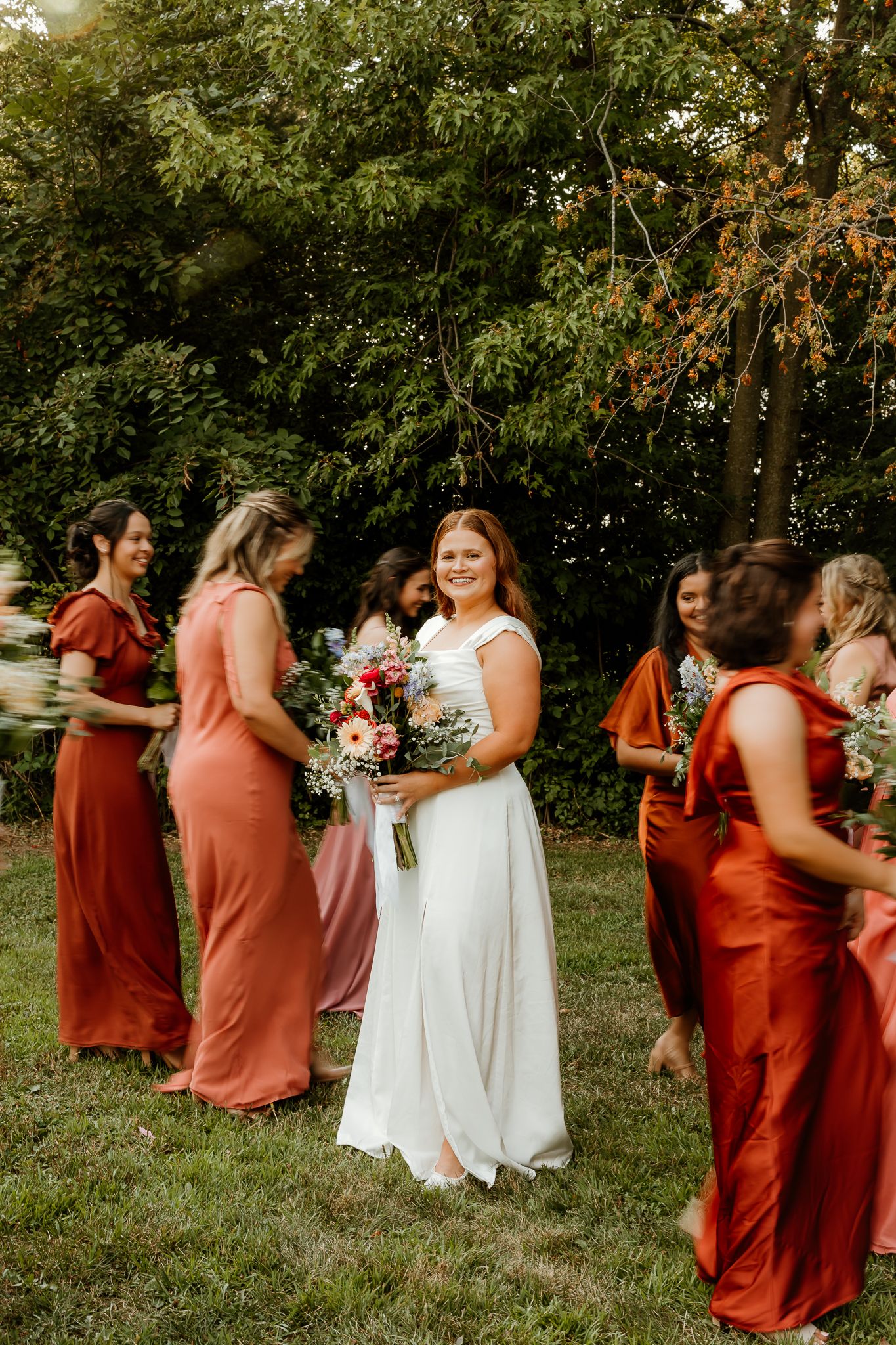 Bride in white gown holds bouquet surrounded by bridesmaids in rust and coral dresses outdoors.