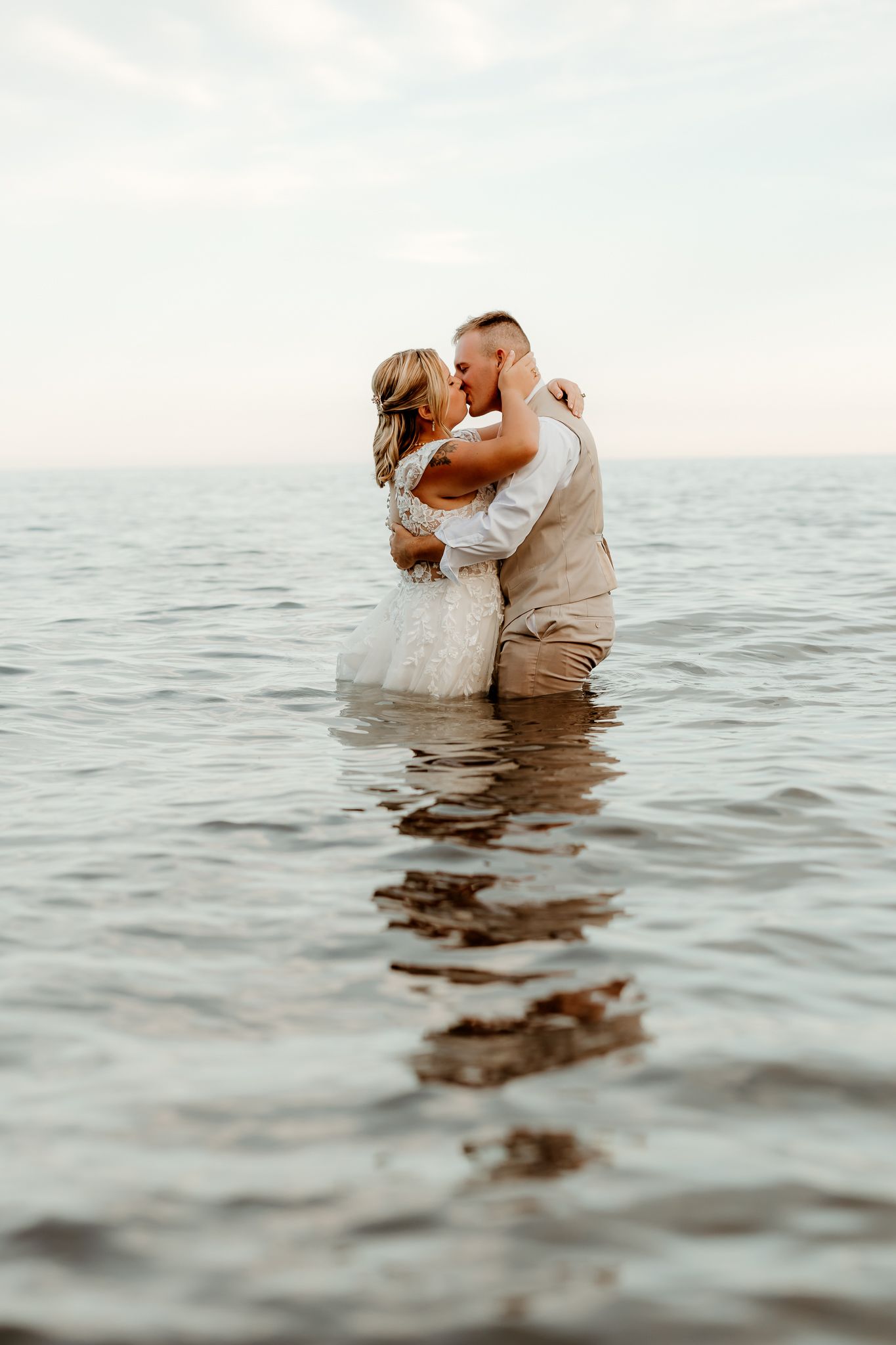 Bride and groom kissing while standing waist-deep in ocean water, wearing wedding attire in a romantic seaside ceremony.