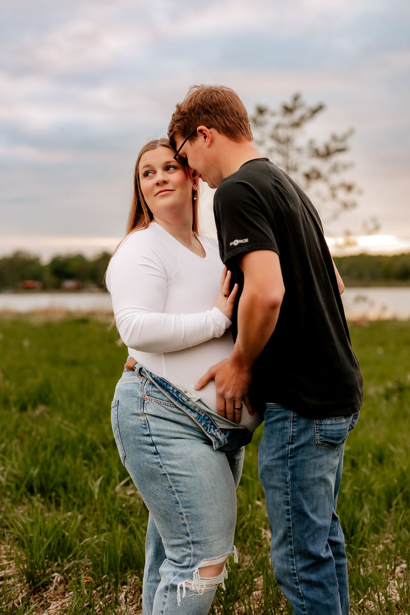 Young couple embracing at sunset in a grassy field, woman in white top and ripped jeans.
