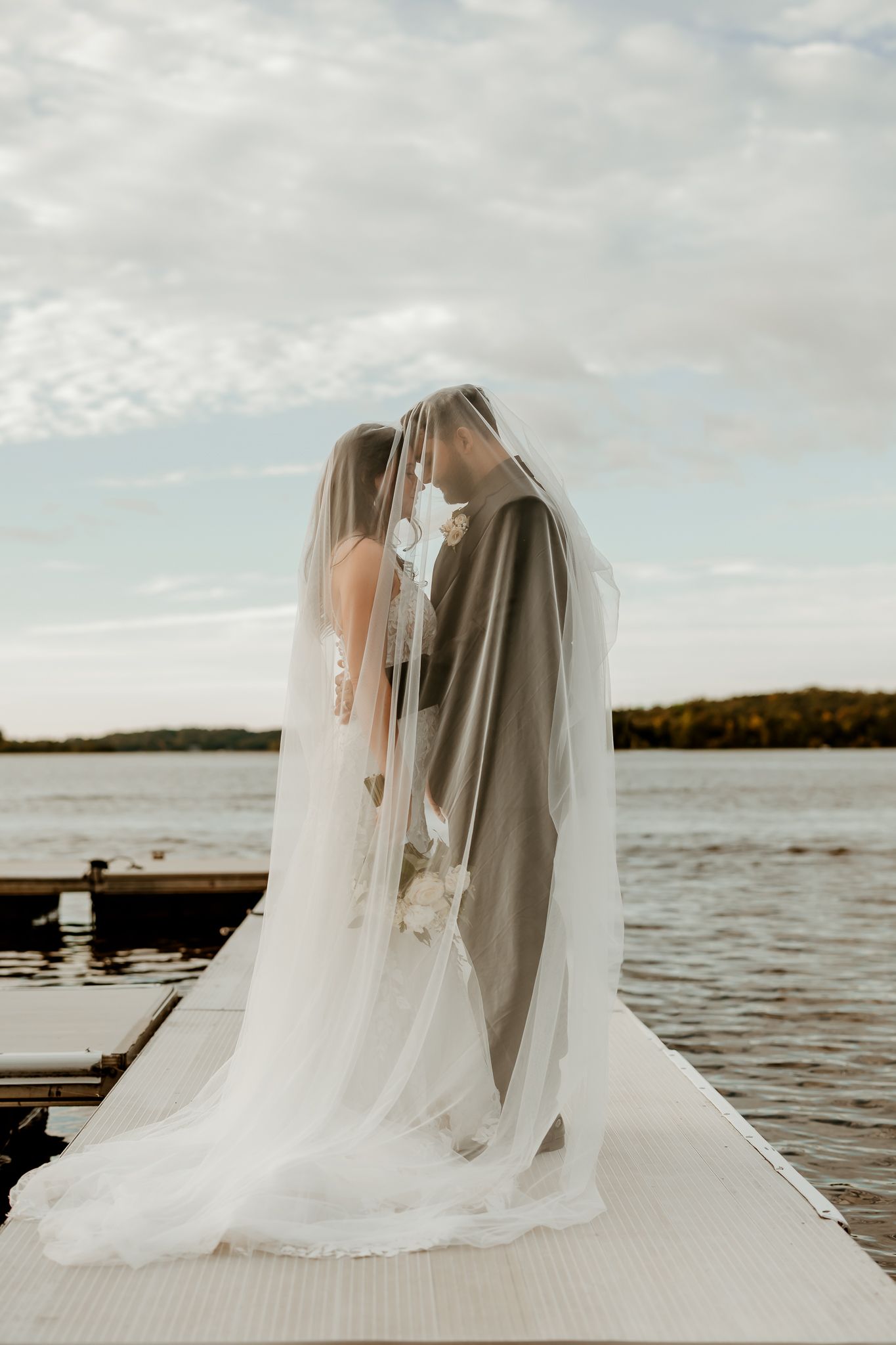 Bride and groom embrace under a sheer wedding veil on a wooden lakeside dock at sunset, with calm water and trees behind them.