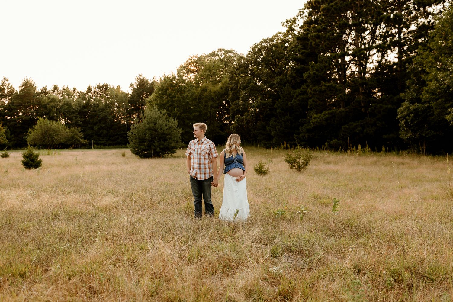 Couple standing in golden field at sunset, pregnant woman in white dress with man in plaid shirt, surrounded by trees.