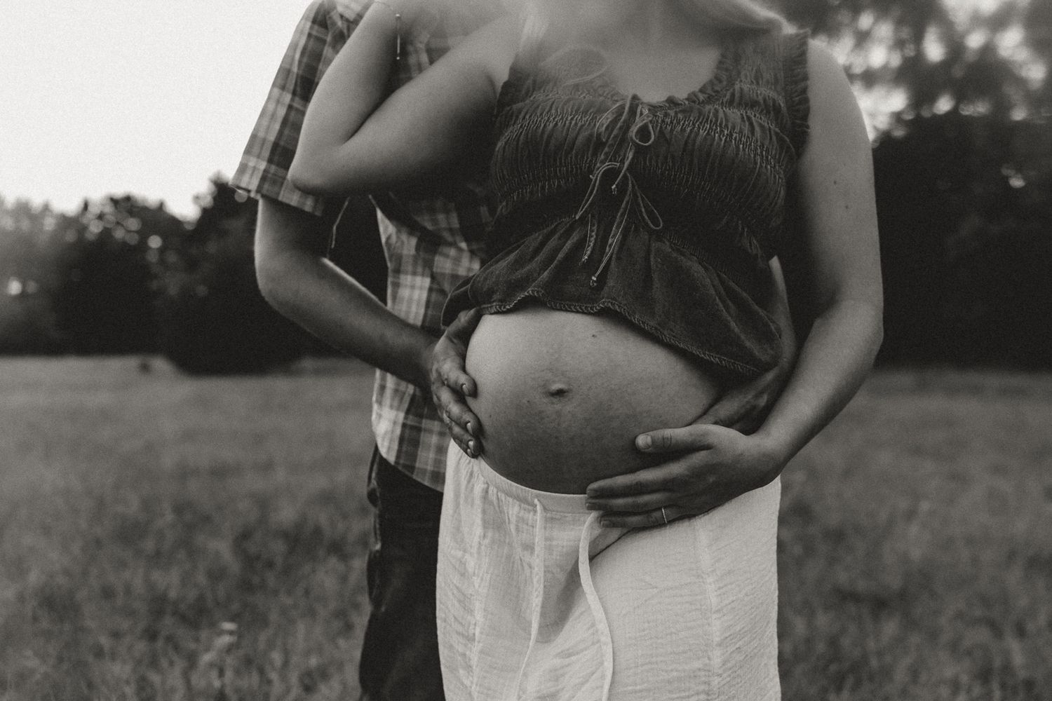 Black and white maternity photo of couple embracing outdoors, showing pregnant belly in field setting.