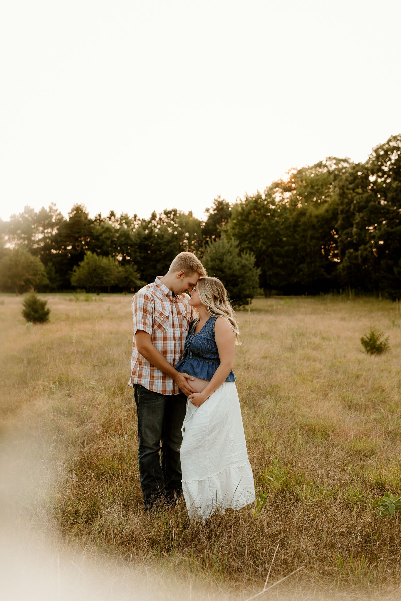 Couple sharing a tender moment in a golden sunlit field, woman in denim top and white skirt, man in plaid shirt.