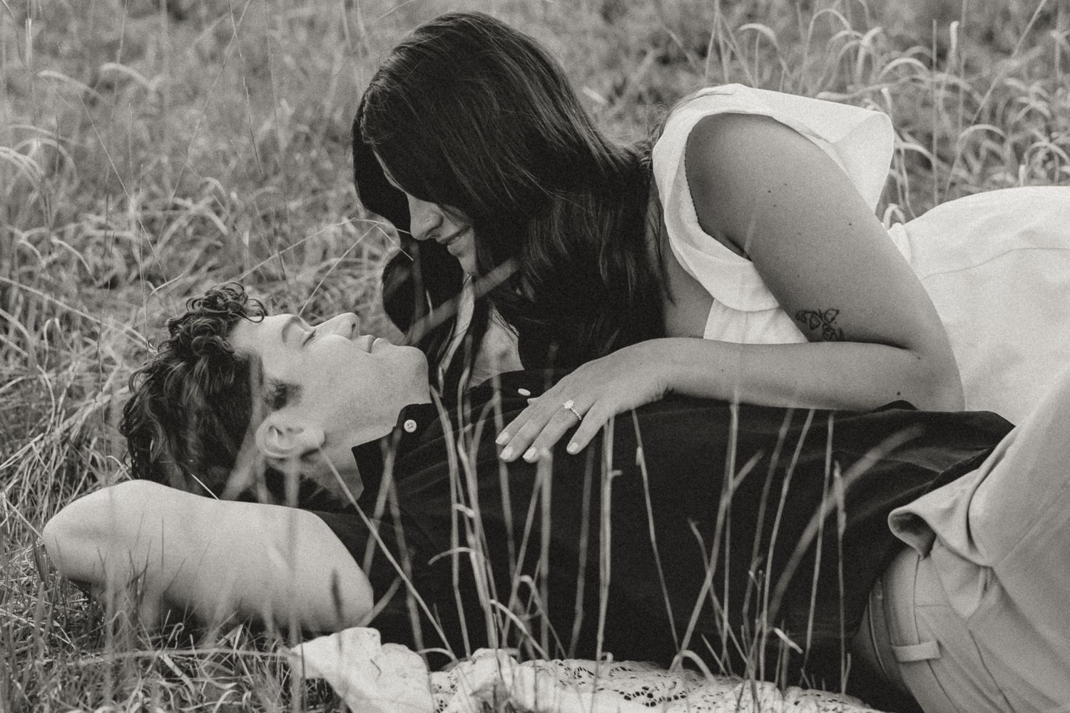 Black and white romantic photo of a couple lying in tall grass, gazing at each other lovingly.