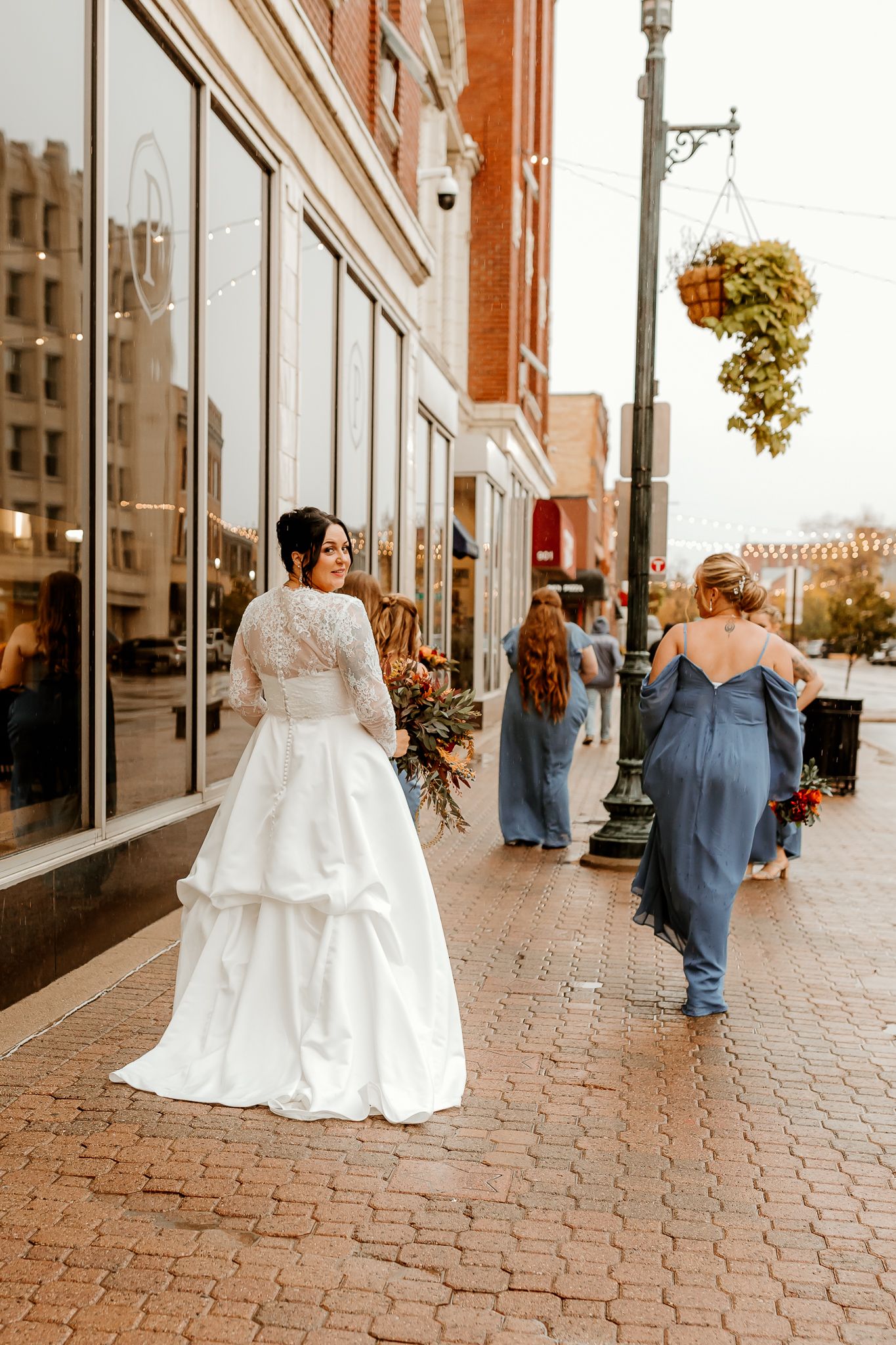 Bride in a white lace-sleeved gown holds a bouquet on a brick sidewalk as bridesmaids in dusty blue dresses walk down a downtown street.