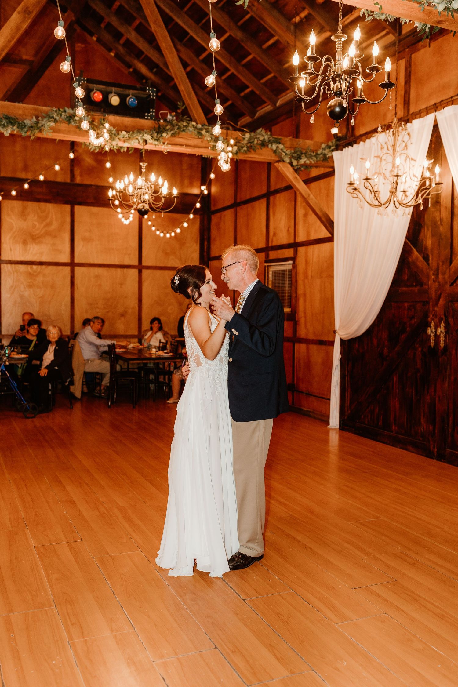 Bride in a white gown and groom in a dark jacket share a slow dance in a rustic wooden barn venue with chandeliers and draped curtains.