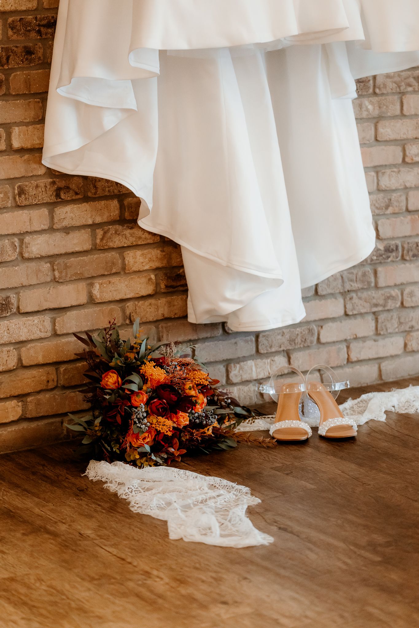 White wedding dress hanging against a brick wall above an orange fall bouquet, lace veil and nude heeled sandals on a wood floor.