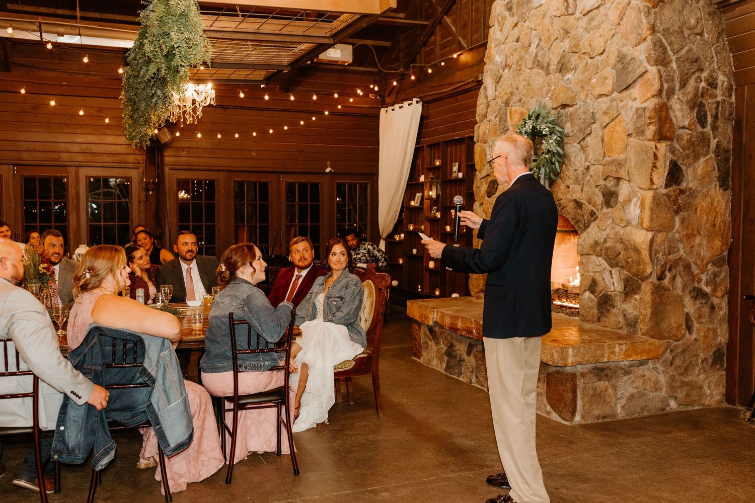 Man in a navy blazer gives a toast to seated wedding guests in a rustic lodge with a stone fireplace and string lights.