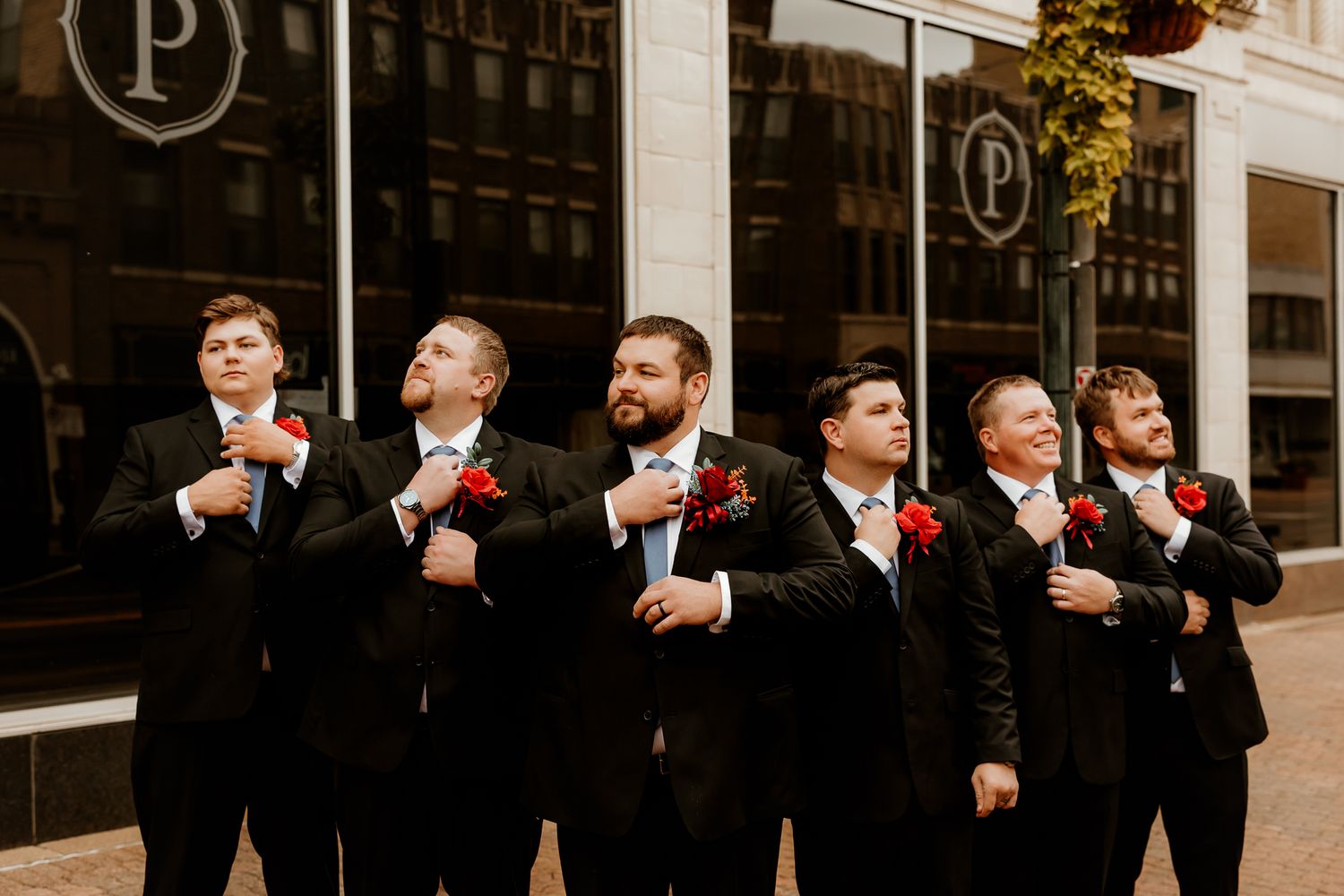 Six groomsmen in black suits with red boutonnieres stand in a row outside a downtown building, adjusting their jackets before a wedding.